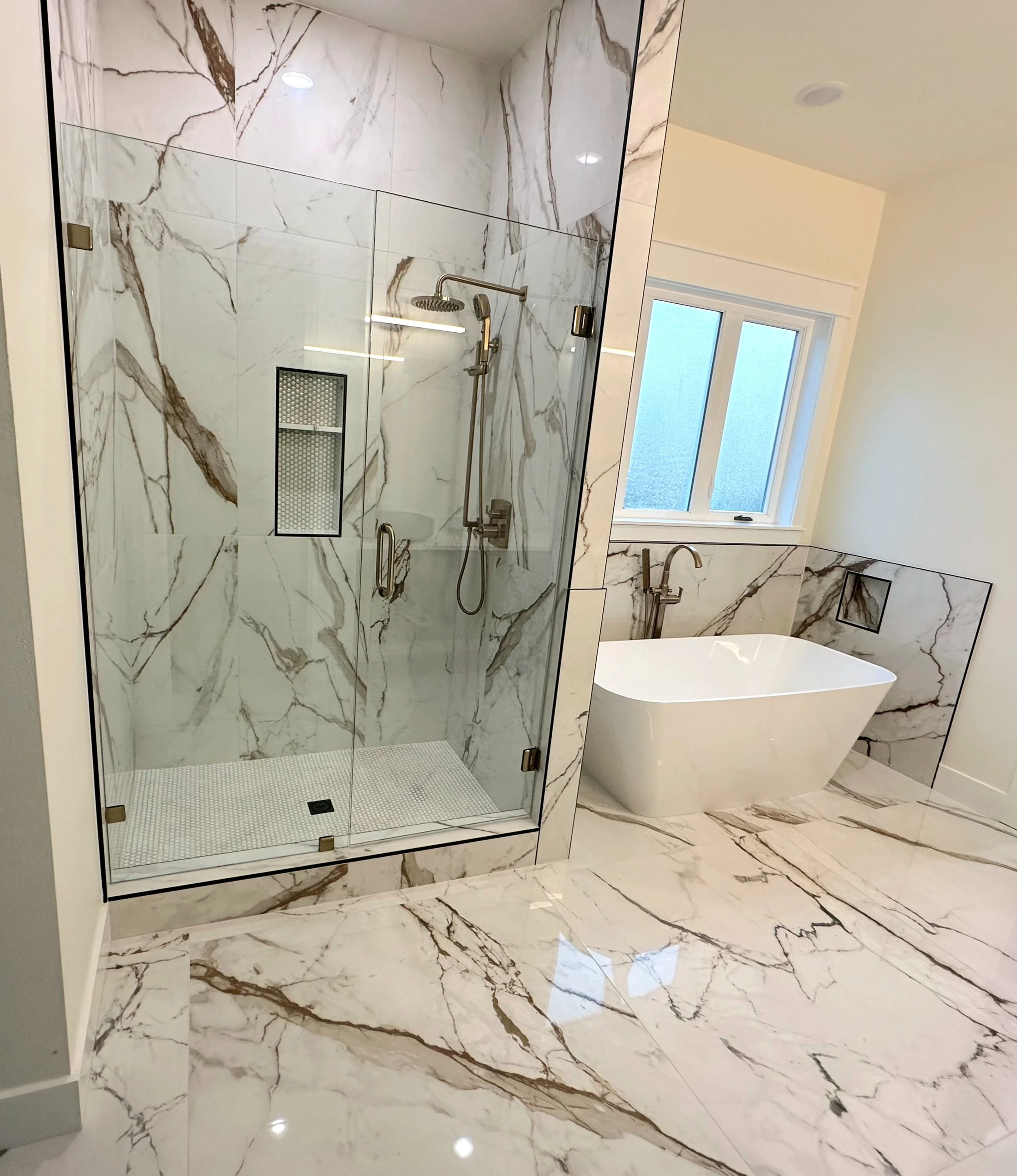 A modern bathroom with a marble shower enclosure, a bathtub, and marble flooring and walls. The shower has glass doors and a metal shower system, with a window allowing natural light into the space.
