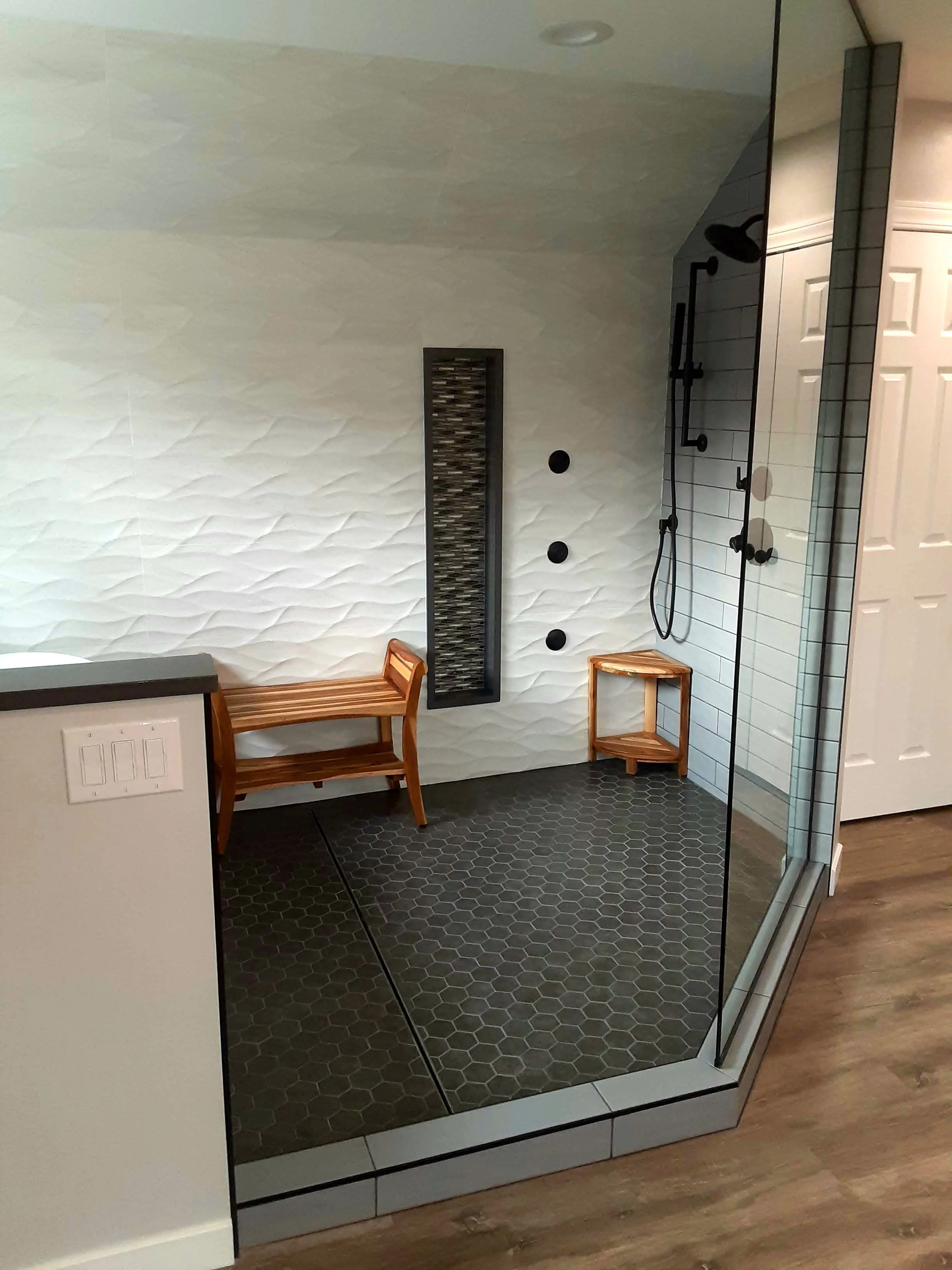 A modern bathroom shower with white textured wall tiles, a black showerhead and fixtures, a narrow vertical decorative panel, and two small round black objects on the wall. There are two small wooden stools and honeycomb-patterned black floor tiles.