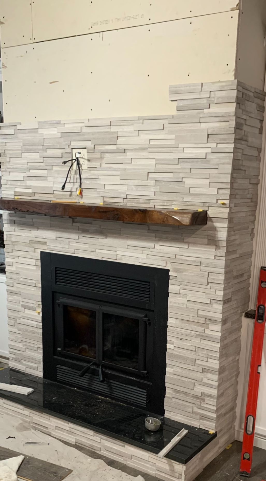 Fireplace with white stone veneer, black metal insert, wooden mantel shelf, and construction tools nearby.