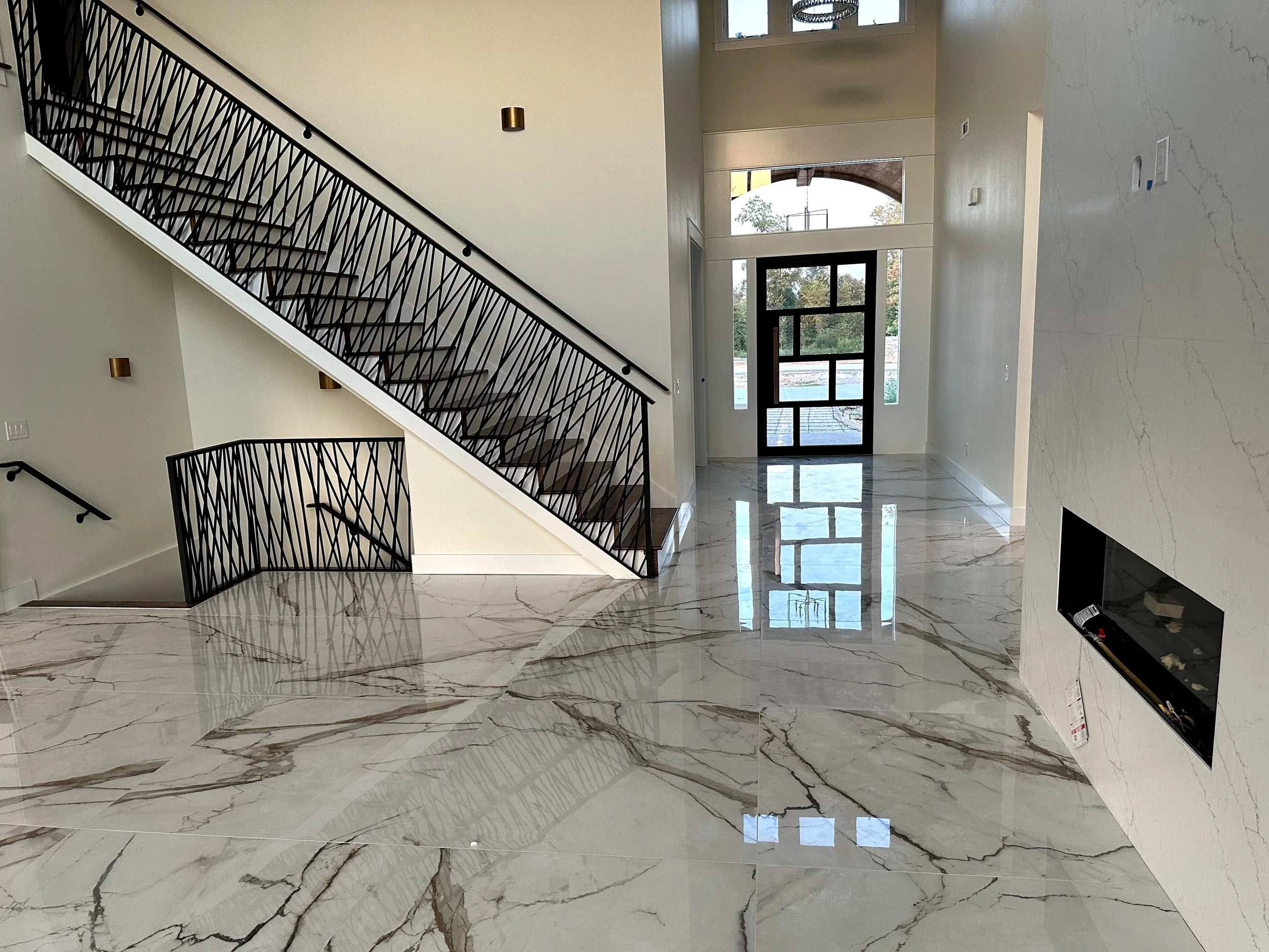 Empty foyer with polished marble floors, a staircase with black metal railings, large windows, and a modern fireplace.