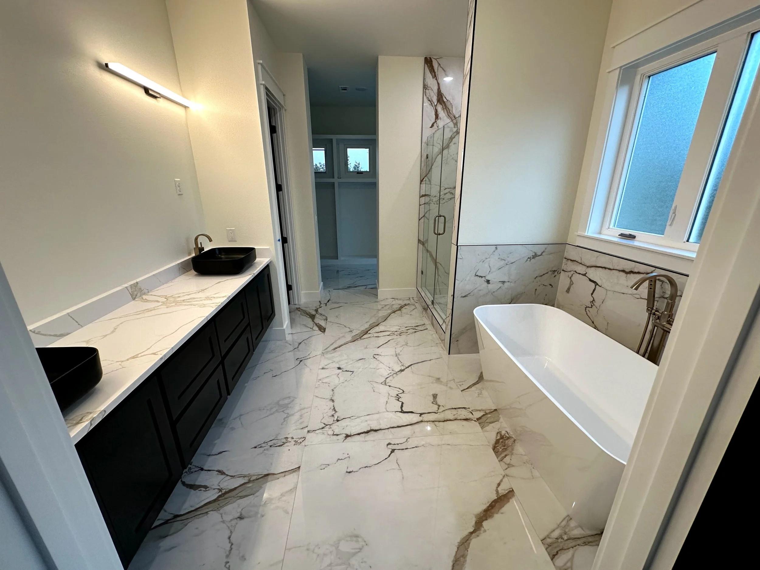 Modern bathroom with marble flooring and walls, a black sink with a gold faucet on a white marble counter, a separate shower with glass doors, a freestanding bathtub, a window, and a wall-mounted light fixture.
