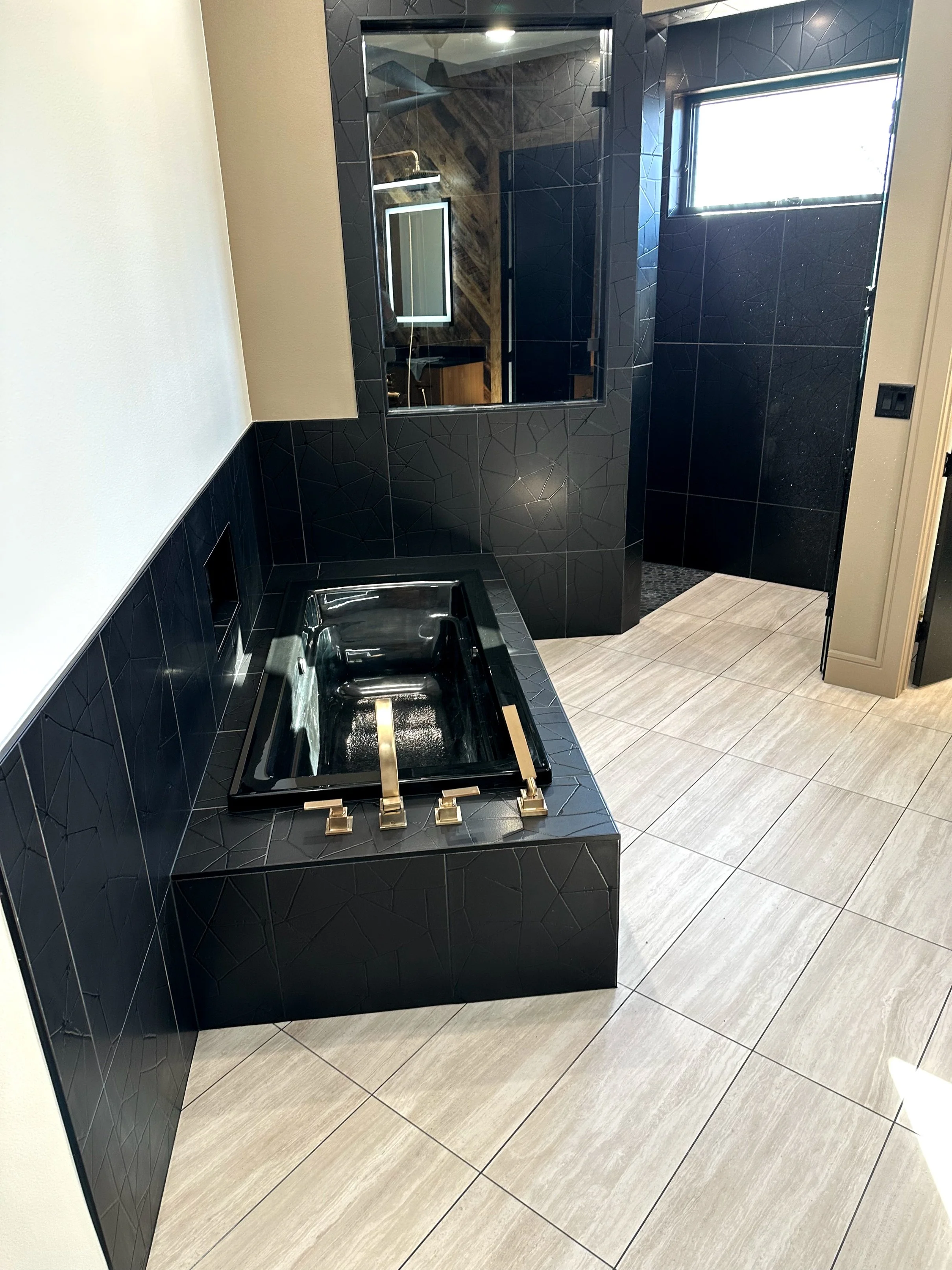 Black vanity with a built-in sink, a black framed mirror, black tile walls, a window, beige floor tiles, and a shower area with a glass door and wooden wall accents.