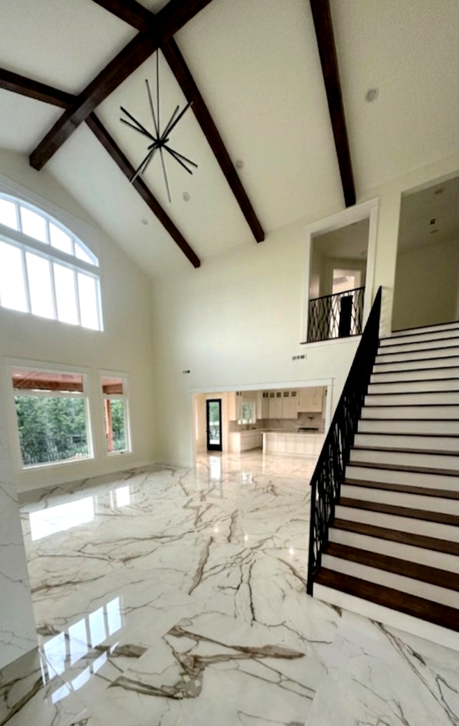 Spacious living area with high vaulted ceiling and dark wood beams, white walls, marble floor, large arched window, and staircase with black railings leading to the upper level, open kitchen visible in the background.