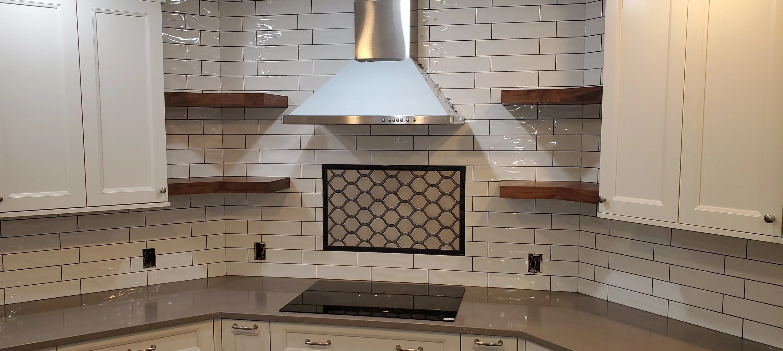 A kitchen wall with white subway tiles, wooden shelves, white cabinetry, a stainless steel range hood, and a decorative vent with a black frame.