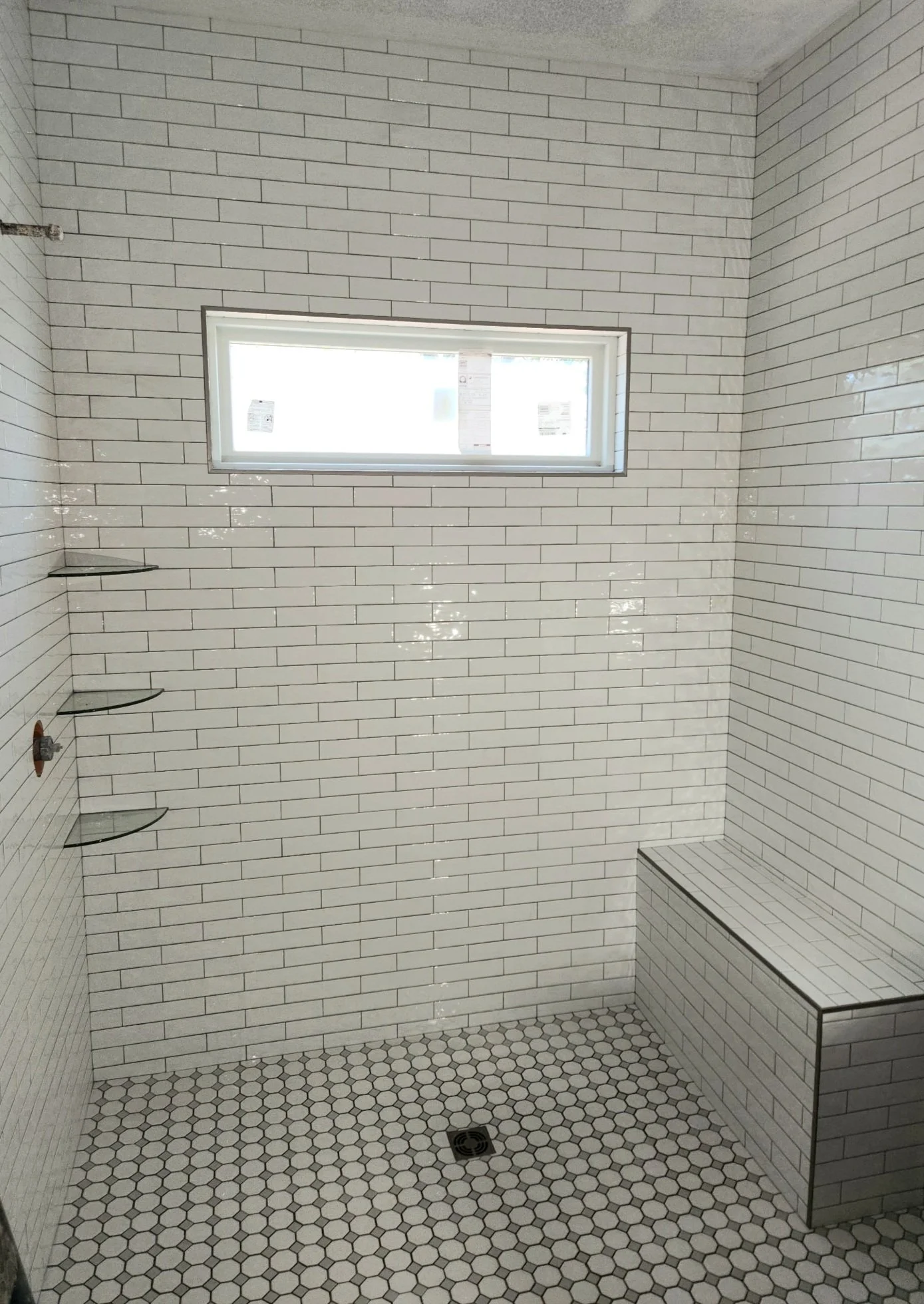 Unfinished shower with white subway tiles on the walls, small hexagon tiles on the floor, a window, a built-in bench, and three small corner shelves.