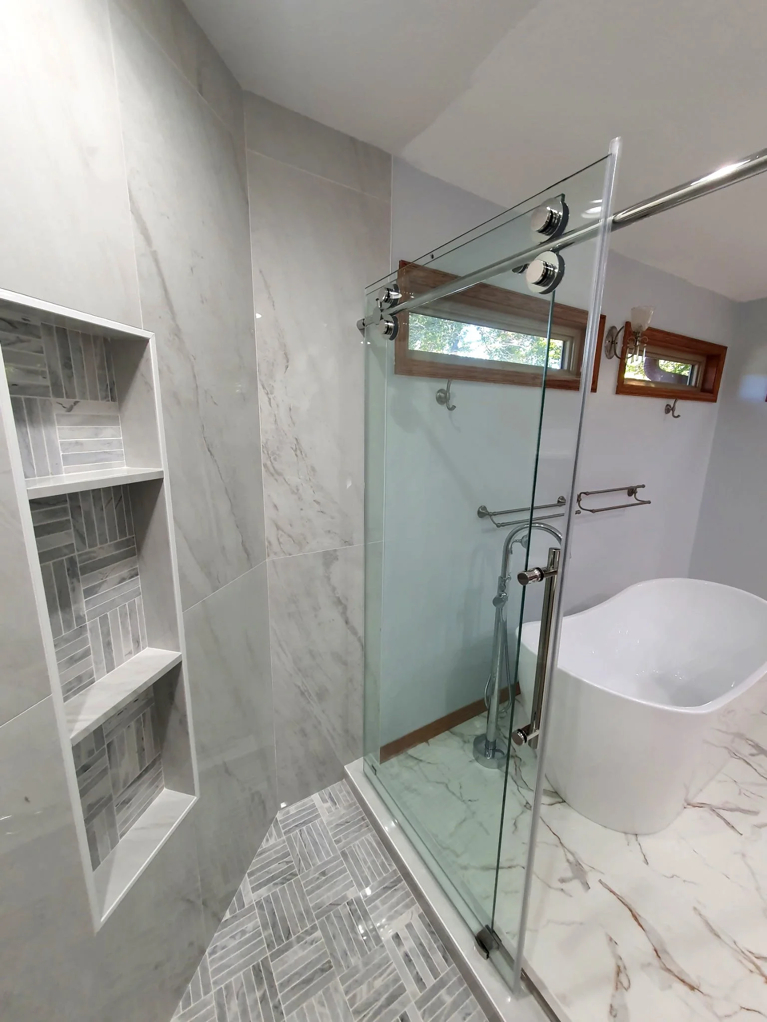 Modern bathroom with marble floor and wall tiles, glass shower enclosure, and a free-standing bathtub. Two small rectangular windows with wooden frames provide natural light.