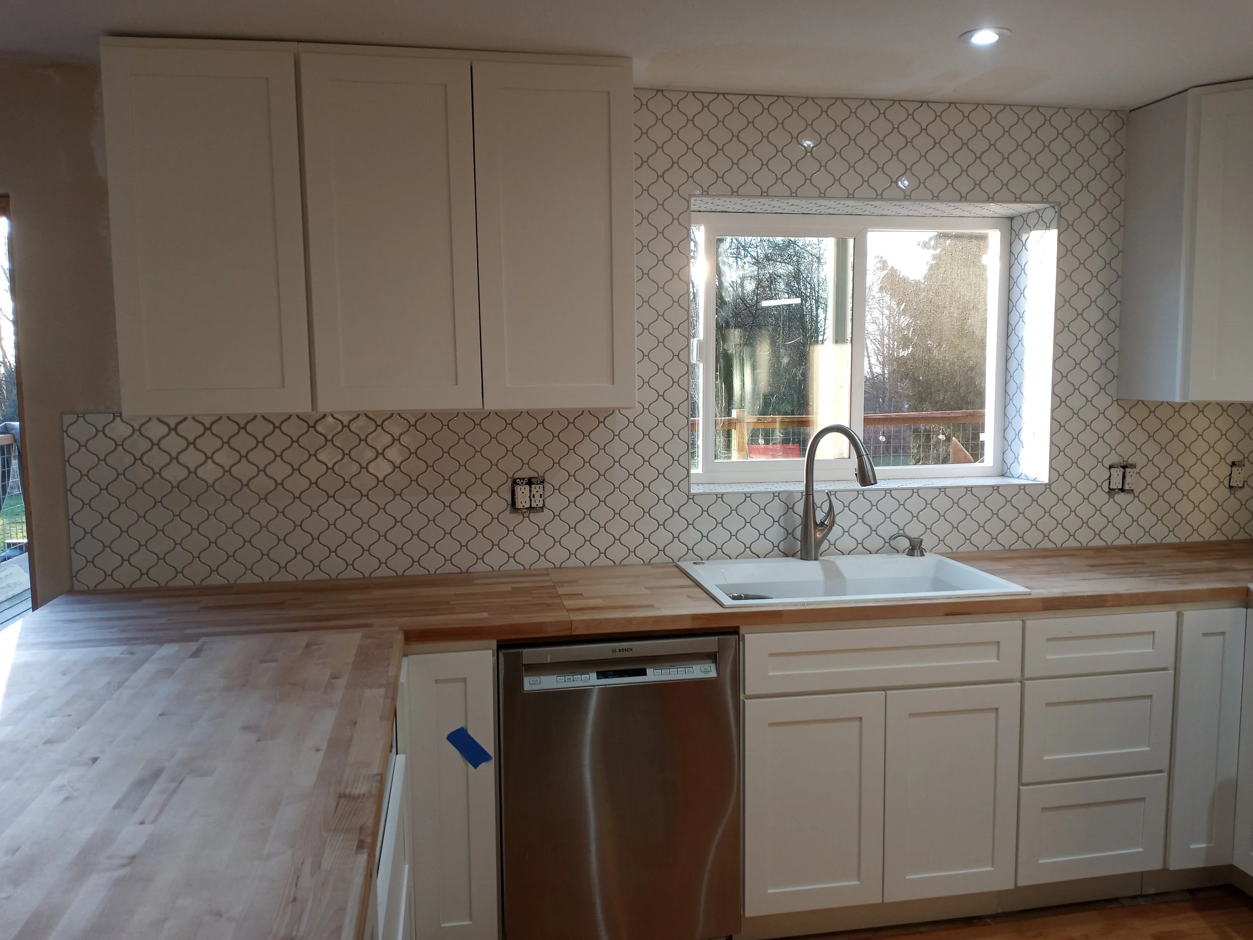 Modern kitchen with white cabinets, a patterned tile backsplash, a large window, a sink with a faucet, and a dishwasher beneath the counter.