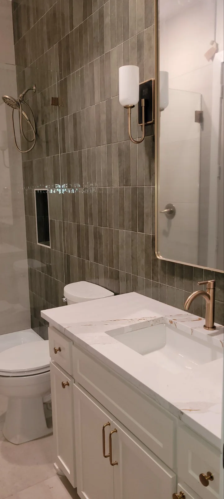 A modern bathroom featuring gray tiled walls, a white vanity with a marble countertop, a white sink, brass fixtures, a large mirror, a wall-mounted light, a toilet, and a walk-in shower with a handheld showerhead.