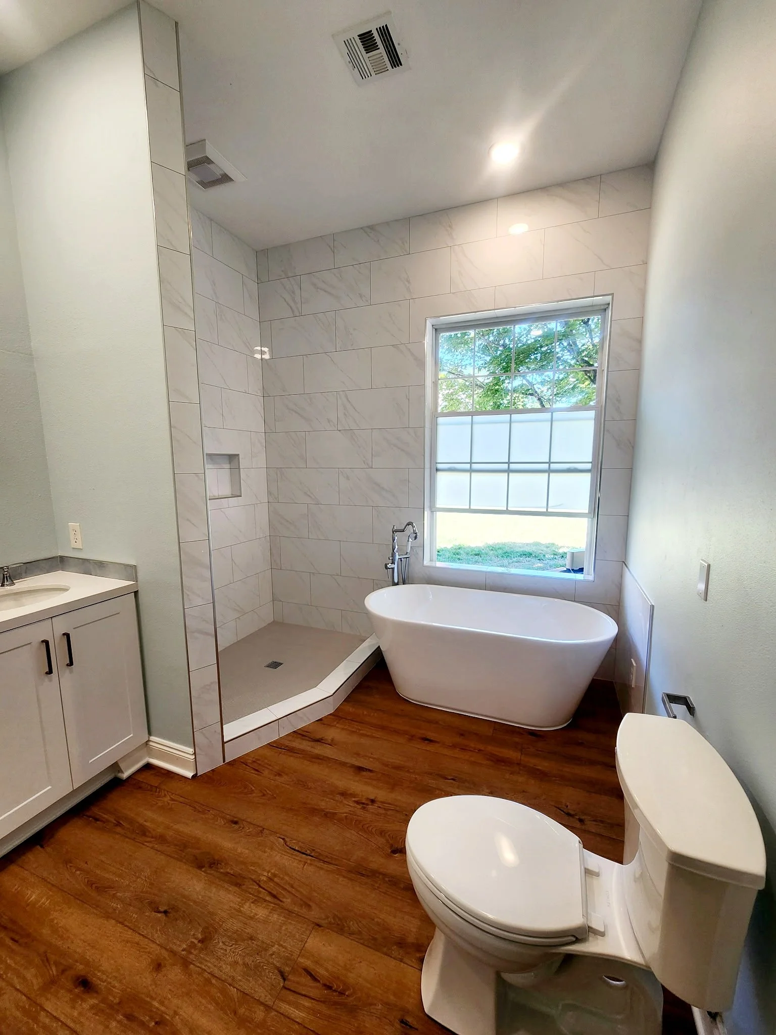 Modern bathroom with a standalone bathtub near a large window, a glass-enclosed shower area, a white vanity with cabinet storage, and a toilet on a wood floor.