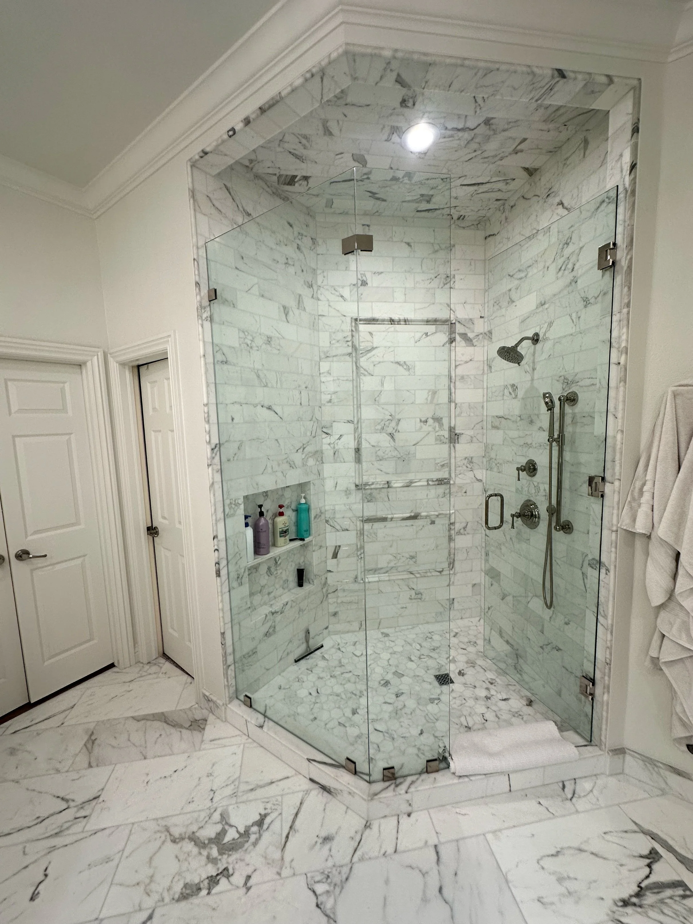 A bathroom with a glass-enclosed shower featuring marble tiles on the walls and floor, equipped with a showerhead, a handheld shower, and bottles of toiletries on a built-in shelf, with white walls and doors nearby.