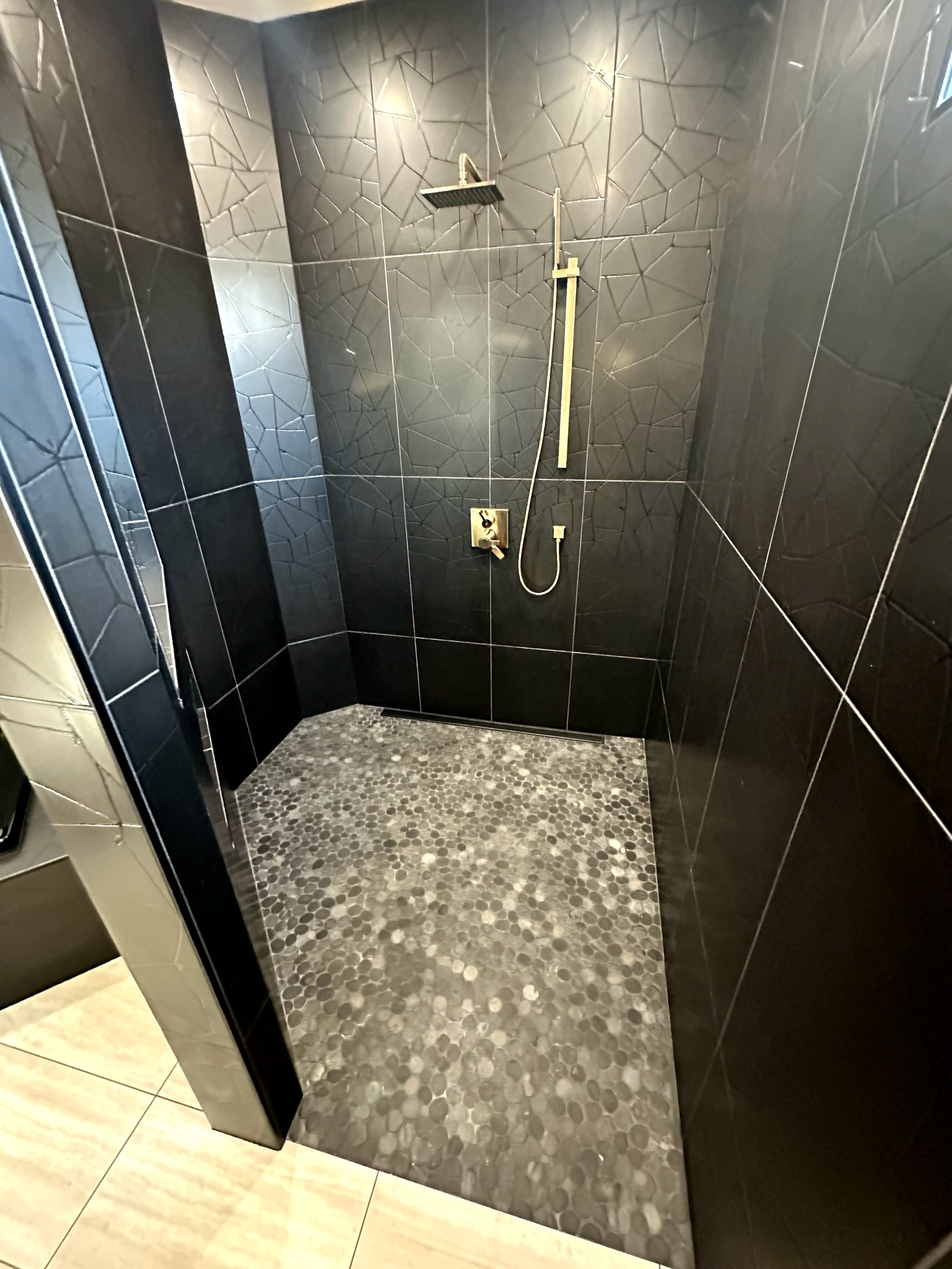 A modern shower with black tiled walls, a rainfall showerhead, a handheld shower wand, and a pebble stone floor.