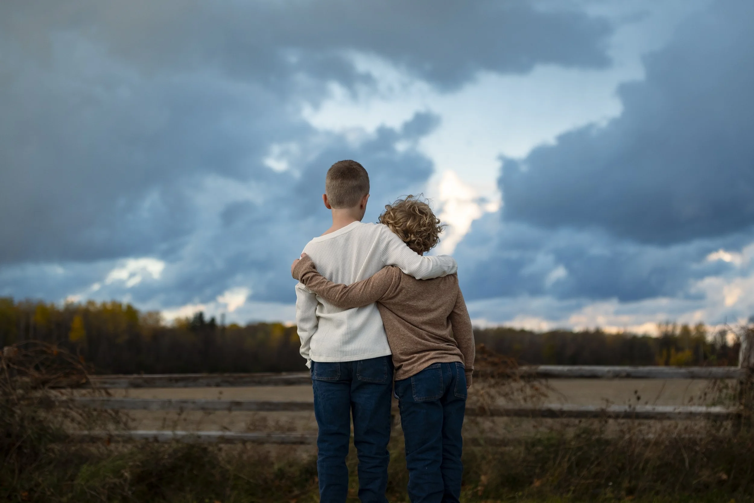Two children standing outdoors, with one child embracing the other, looking at a cloudy sky in the distance.