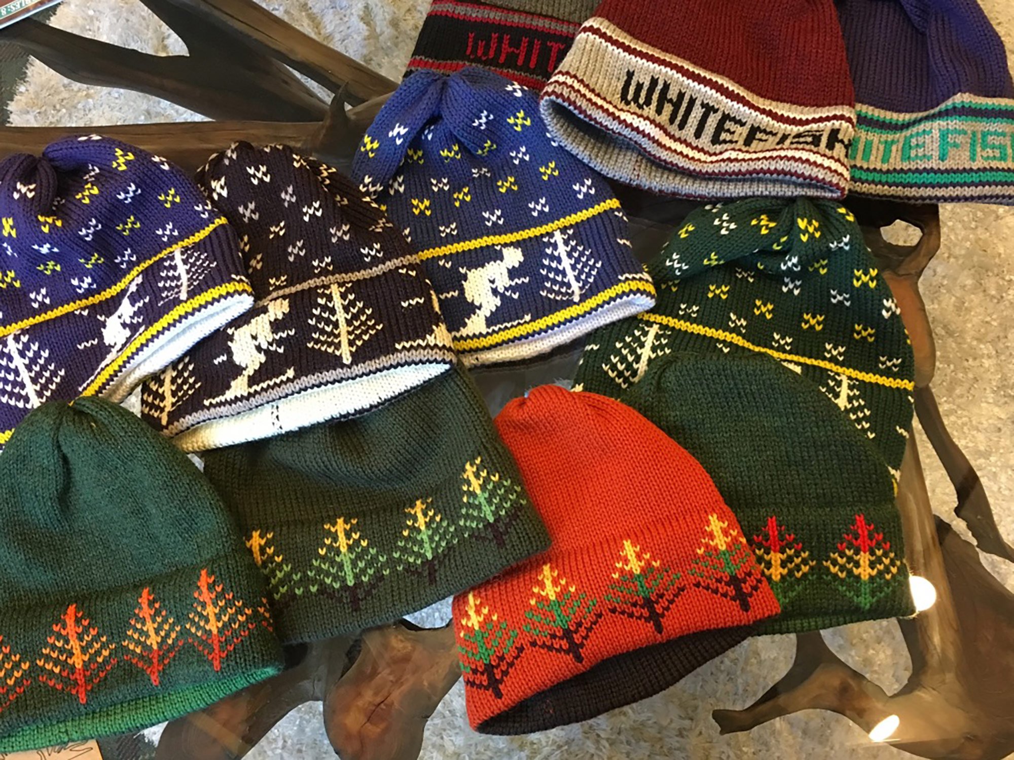 Hand-knit wool beanies with tree and deer patterns, some with 'Whitefish' text