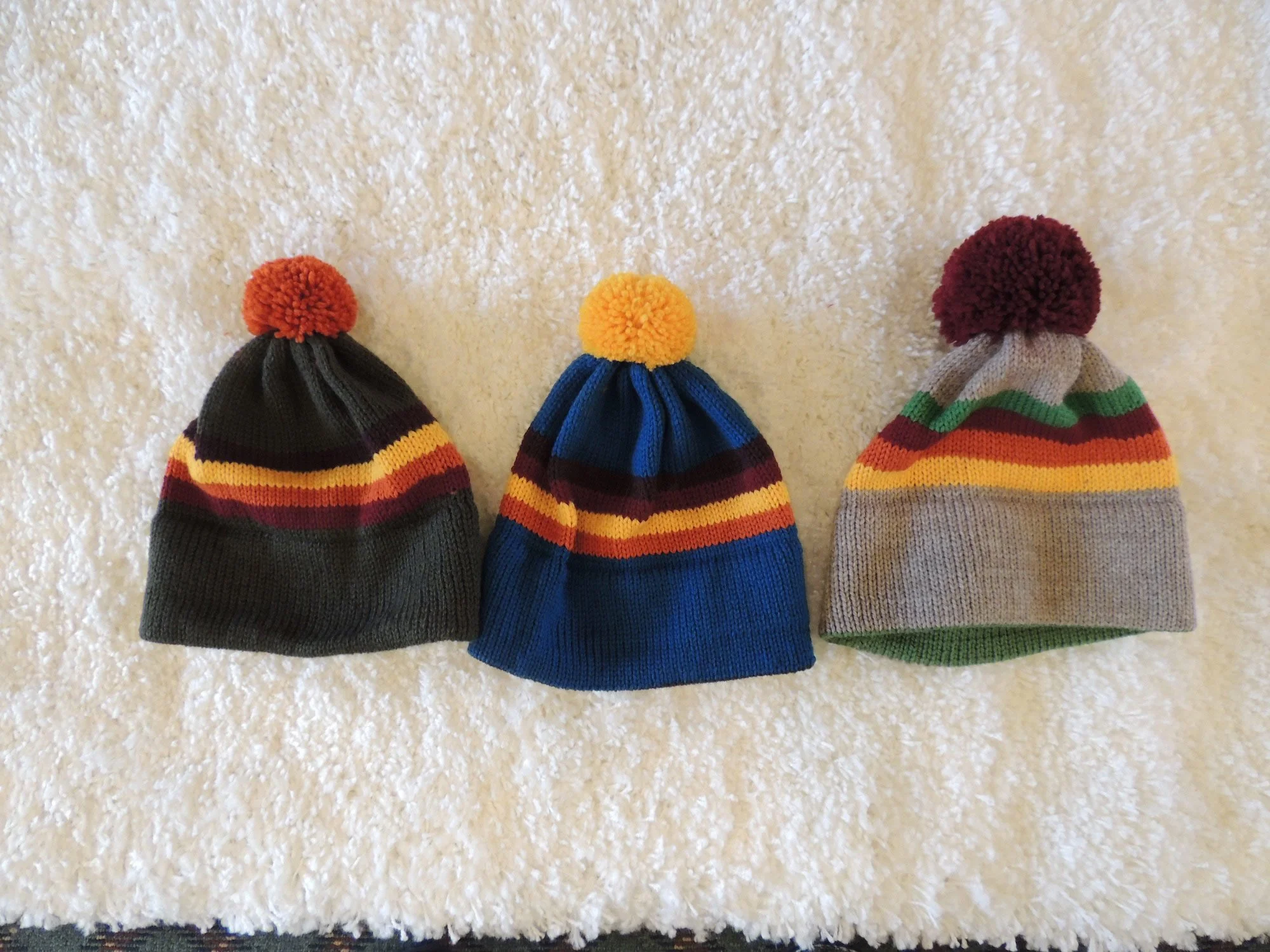 Three hand-knit striped beanies with pom-poms in different colorways