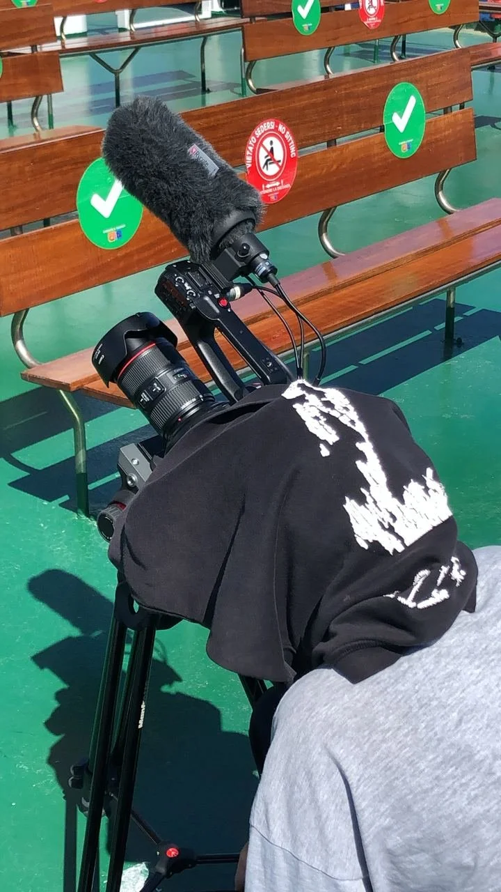A person with a hoodie over their head is operating a camera on a tripod, recording or photographing a scene on a green-colored floor. The scene includes a wooden bench with social distancing markers and signs.