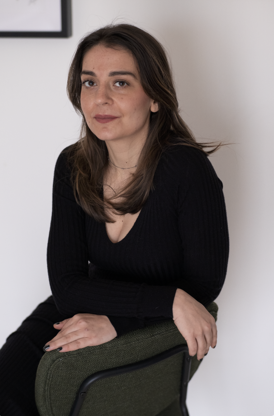 A woman with brown hair wearing a black long sleeve top, sitting on a green chair, with a small black necklace, in front of a white wall.
