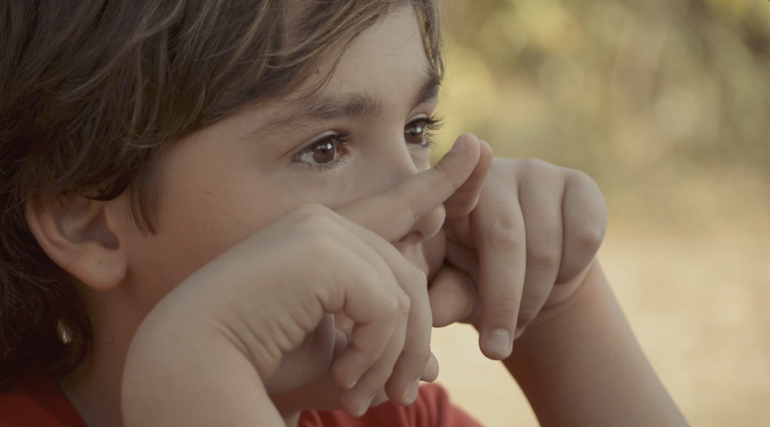 Close-up of a young girl with brown hair and brown eyes, holding her hands near her face, outdoors with yellowish blurred background.