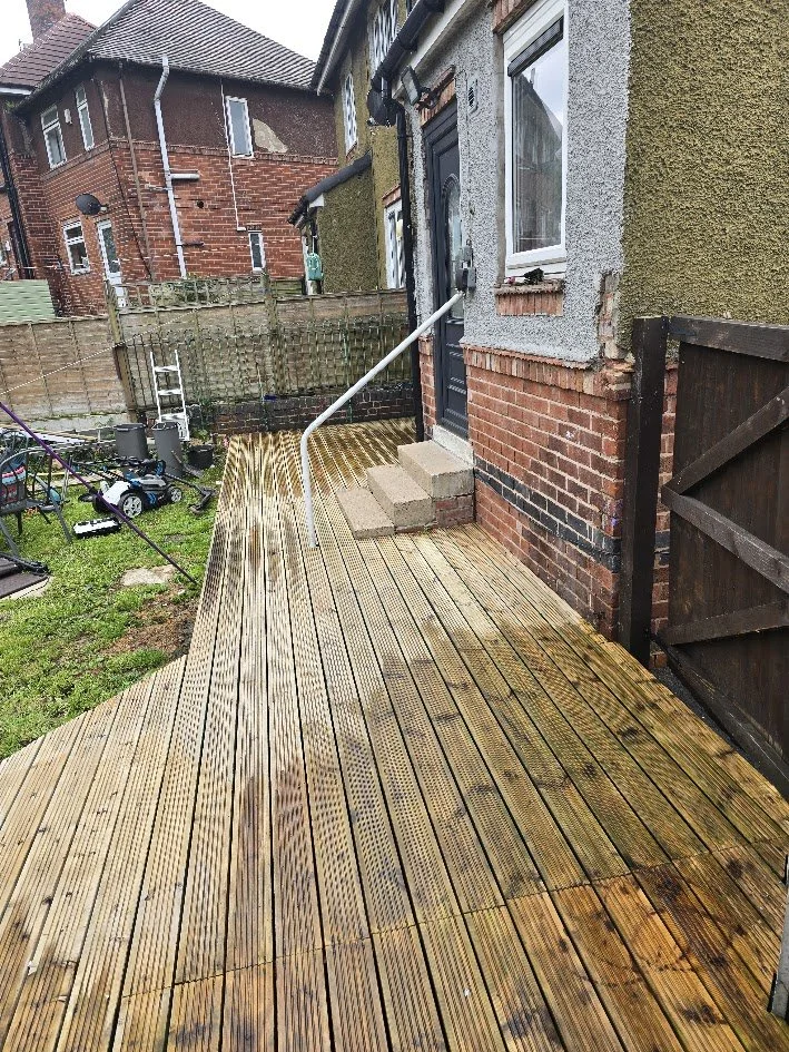 Newly installed wooden deck outside a brick house with steps leading to a black door, surrounded by a backyard with lawn, fencing, and garden equipment.