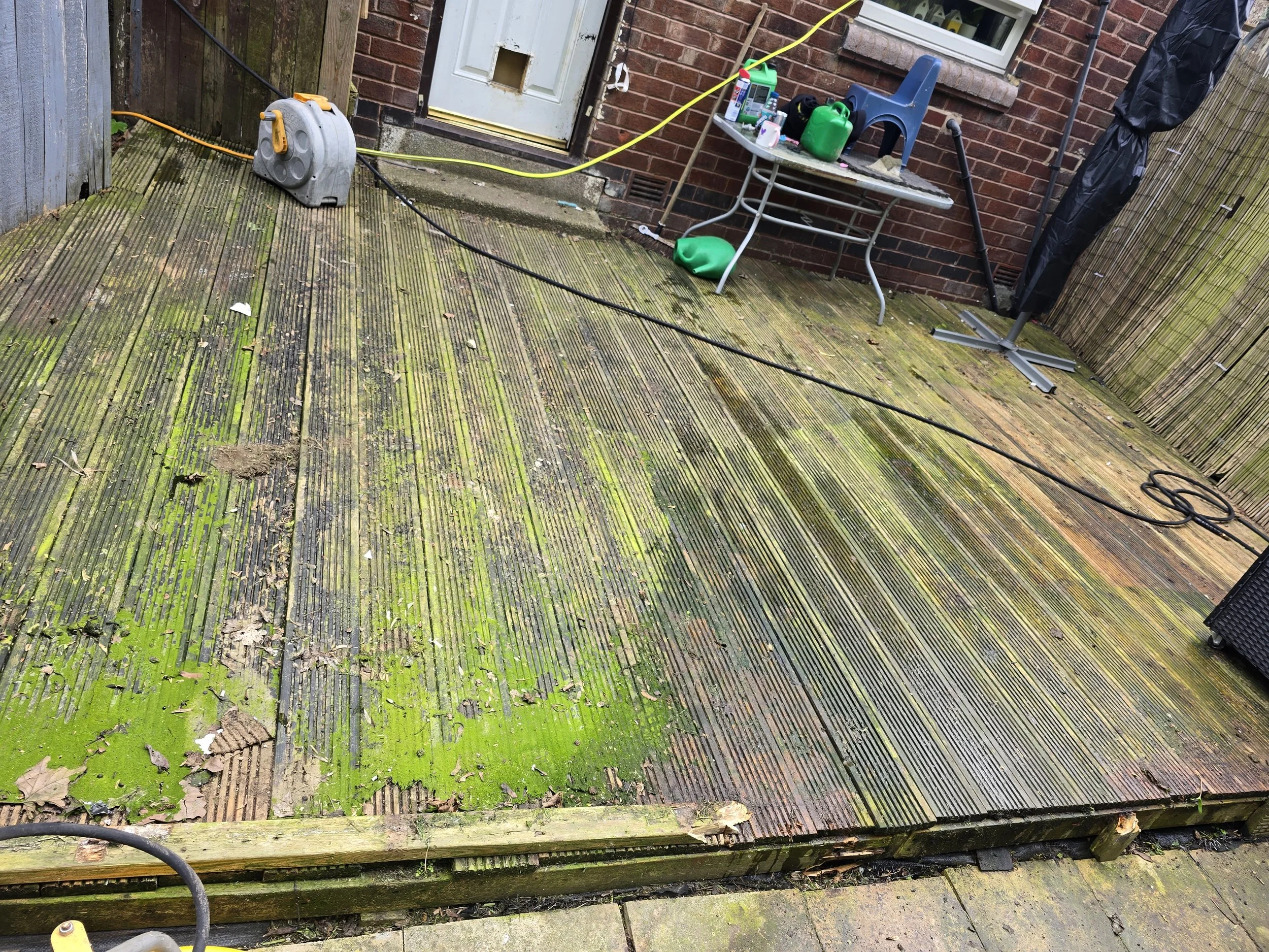A wooden deck with green algae, leaves, and dirt. There is a white door on a brick house with some outdoor items on a table, including a green water container, a black fireplace, and a red and white bottle. Electrical cords and a power reel are on th