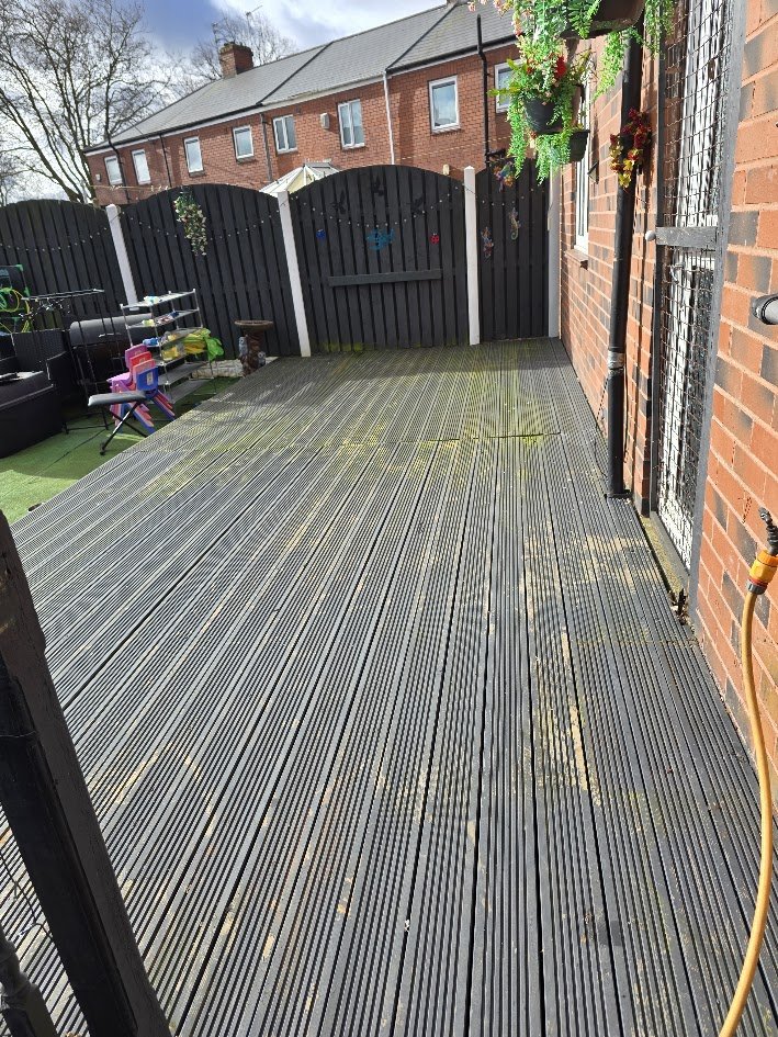 A backyard deck with a black wooden fence and potted plants hanging on the brick wall. There are children's toys, a bench, and cleaning supplies on the deck.