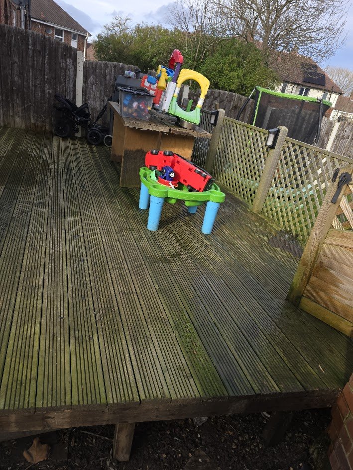 Backyard deck with children's toys, plastic trucks, and a black stroller coffee table, surrounded by a wooden fence and houses in the background.