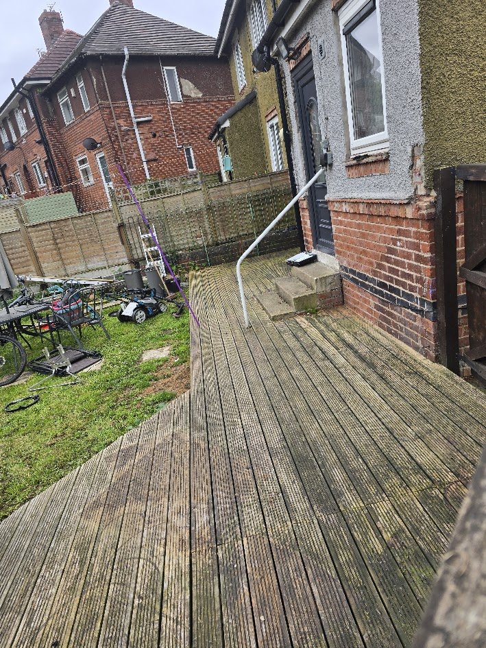A backyard wooden deck attached to a brick house, with a small set of concrete steps leading to a black front door. There is a railing on the deck, and various objects including children's toys, chairs, and gardening tools scattered around the yard.