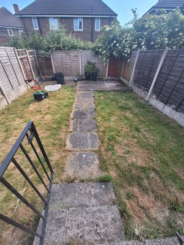 View of a backyard with a stone pathway leading to a small patio area with two large potted plants, fencing on all sides, and a house in the background. There are lawn chairs and gardening tools nearby.