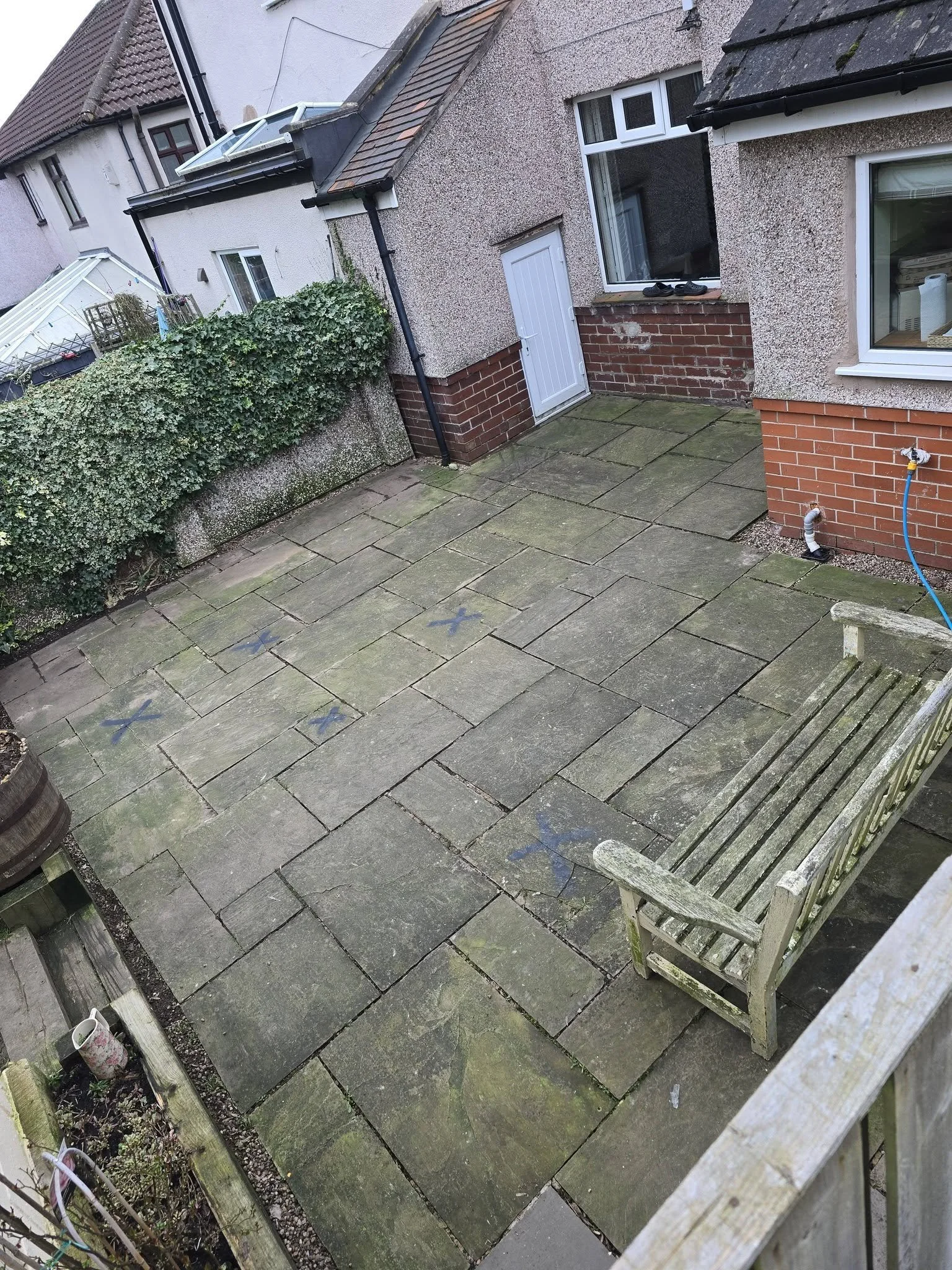 Backyard patio with large square stone tiles, a weathered wooden bench, a small wall with a garden, and neighboring houses with textured walls and shingled roofs.