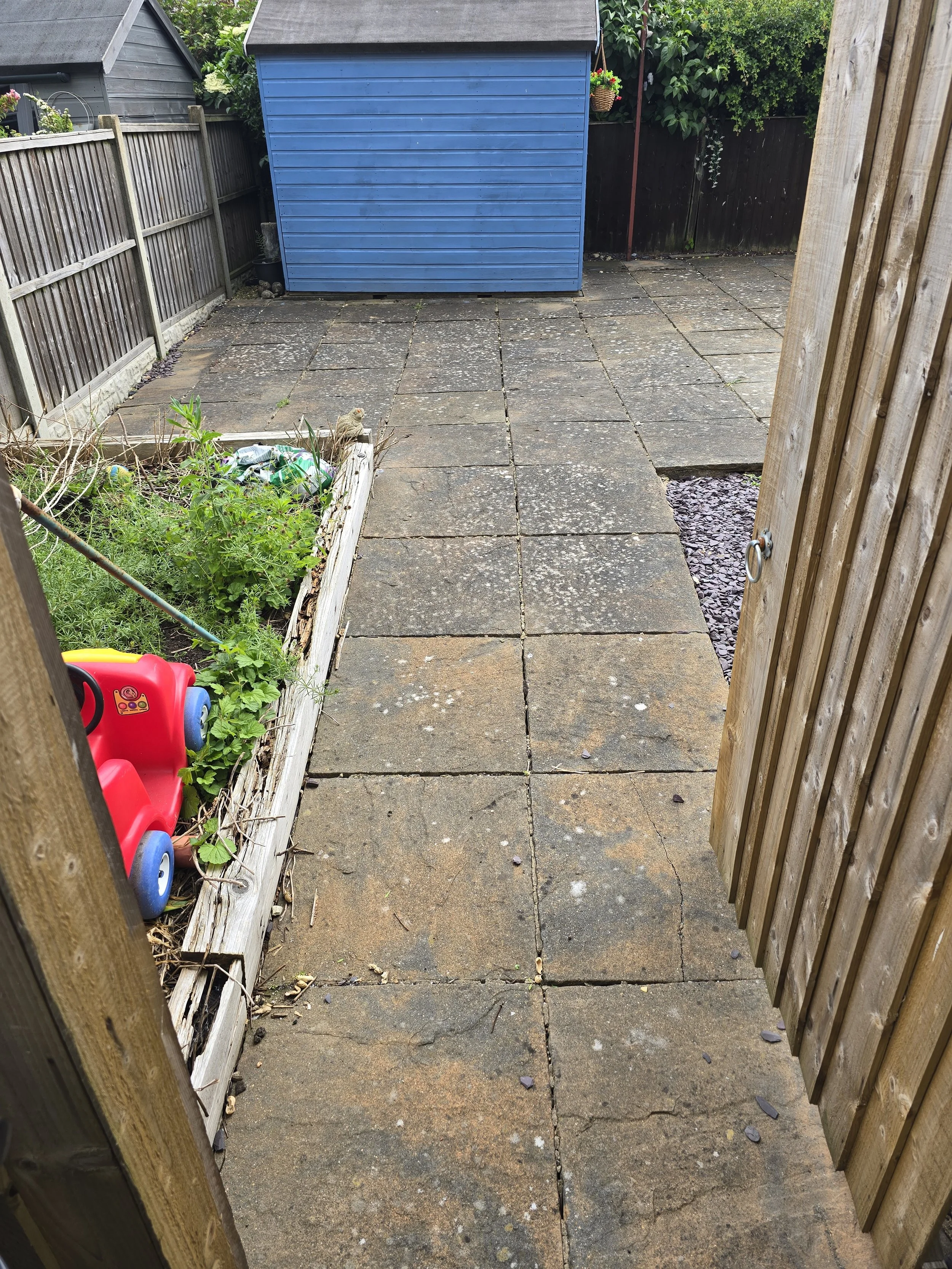 View of a backyard patio seen through an open wooden gate, with a blue shed in the background, a small garden bed on the left with gardening tools and a toy car, and a black fence with greenery behind.