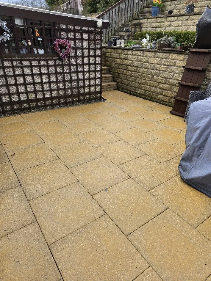 An outdoor patio area with tan paver stones, a wooden staircase leading up to a garden with potted plants, and a lattice fence decorated with a pink heart-shaped wreath.