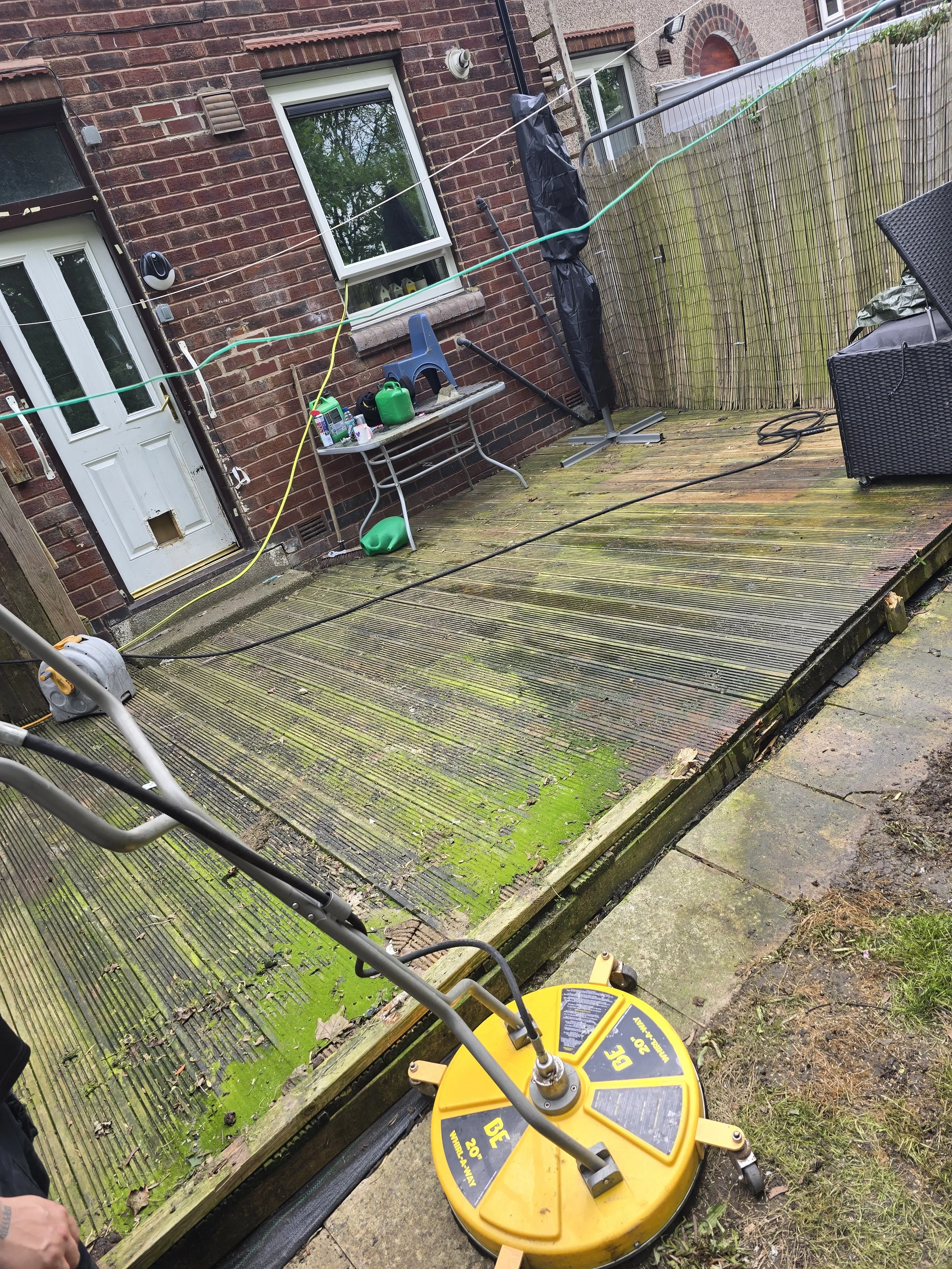 Backyard with wooden deck, brick house wall, window, door with cat door, outdoor table with gardening supplies, garden hose, and pressure washer.