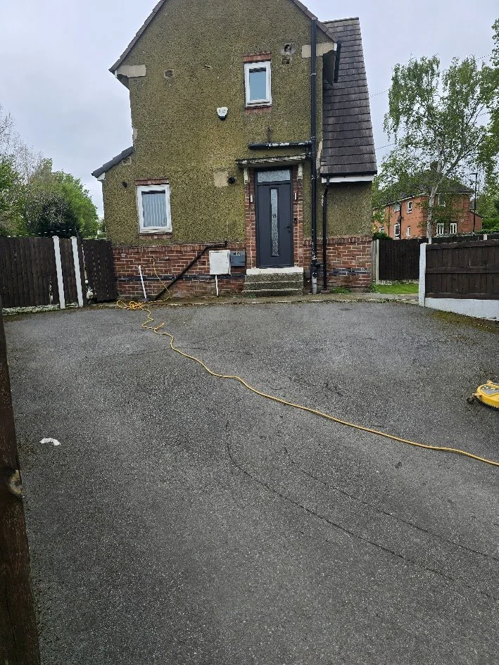 A residential house with a moss-covered exterior, a black front door with a glass panel, a small set of concrete stairs, and a paved driveway with a yellow extension cord and a device on the ground. There are trees and other houses visible in the bac
