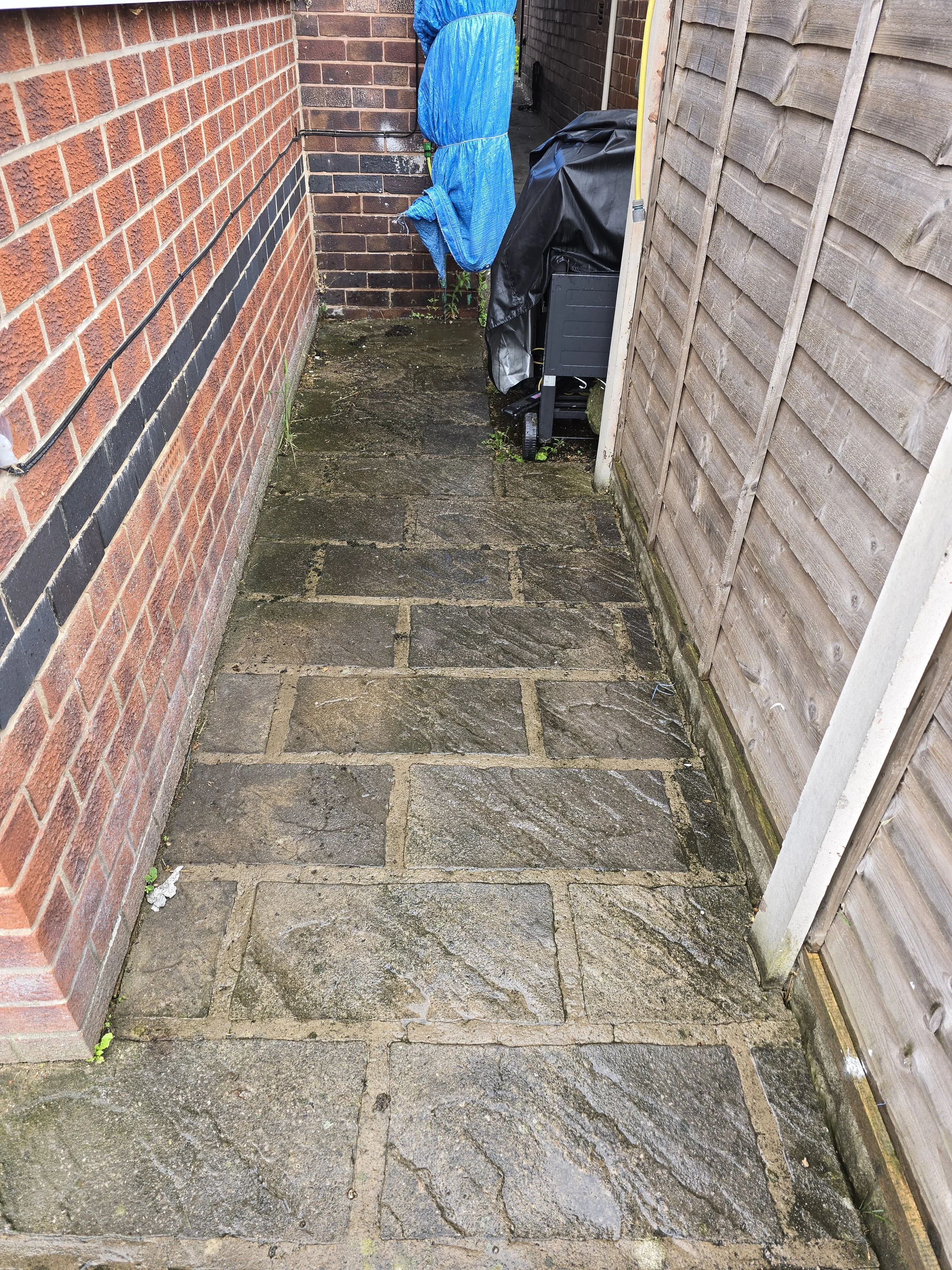 A narrow outdoor area with a red brick wall on the left, a wooden fence on the right, and a wet stone-paved ground. Some objects, including a covered item and a black bag, are positioned near the back corner.