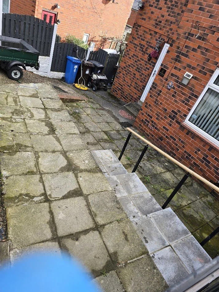 View of a backyard with a small staircase, brick house wall, and various items like trash bins, a wheelbarrow, and garden tools.