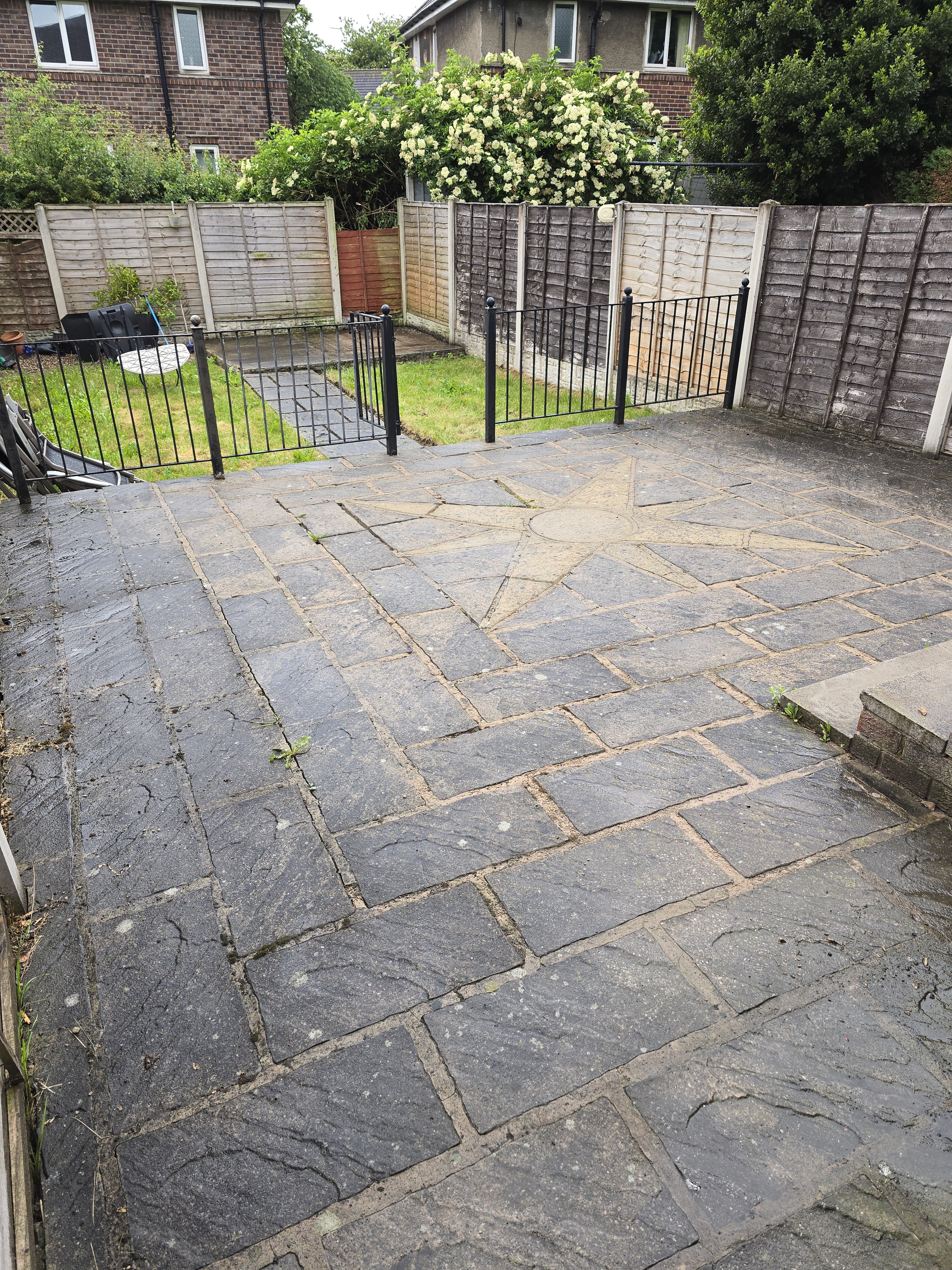A backyard patio with a stone flooring area in the foreground and a grassy lawn area with a small fence and gate. The background shows wooden and brick fences, trees, and houses with windows. There is a garden hose reel, some gardening tools, and a s