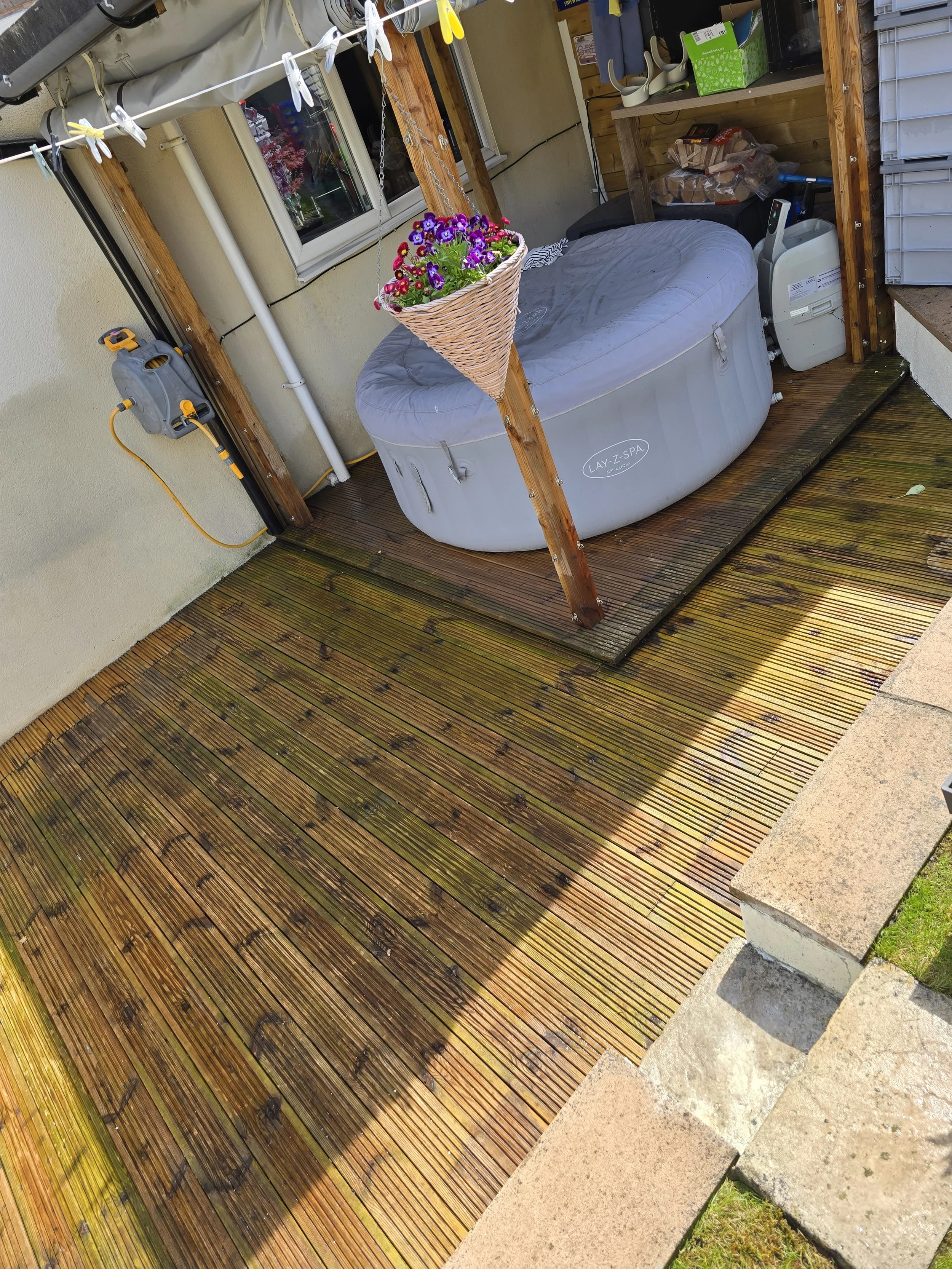 An outdoor wooden deck with a hot tub, a hanging flower basket with purple and pink flowers, a window, and various items on a wooden shelf, including a box of snacks and a small machine, with a bench nearby.