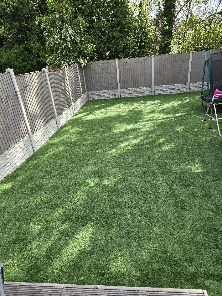 A backyard with well-maintained artificial grass and a gray fence, with some trees and green leaves in the background. There is a pink and purple hammock on the right side.