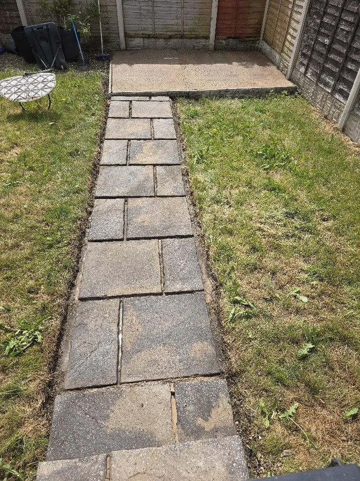 A backyard with a new paved stone pathway leading to a small patio area, with grass on both sides and a wooden fence in the background.