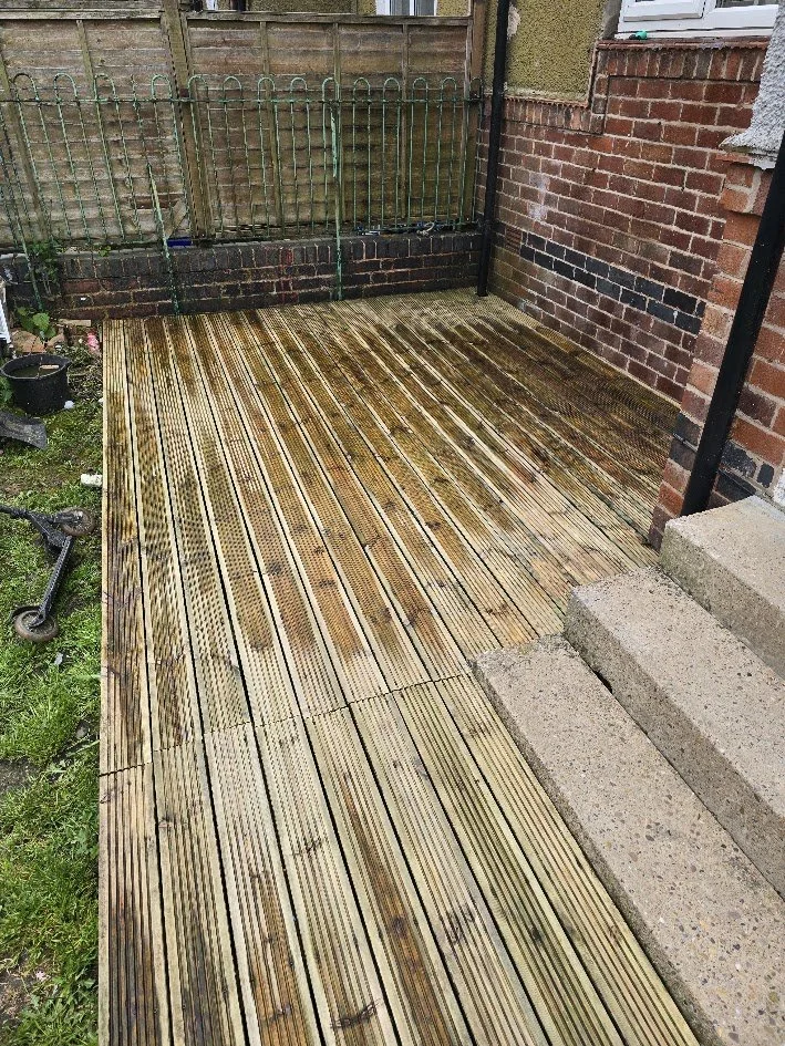 Partially renovated backyard with new wooden decking and concrete steps leading up from the yard, brick fence and wall, with some gardening tools and potted plants visible.