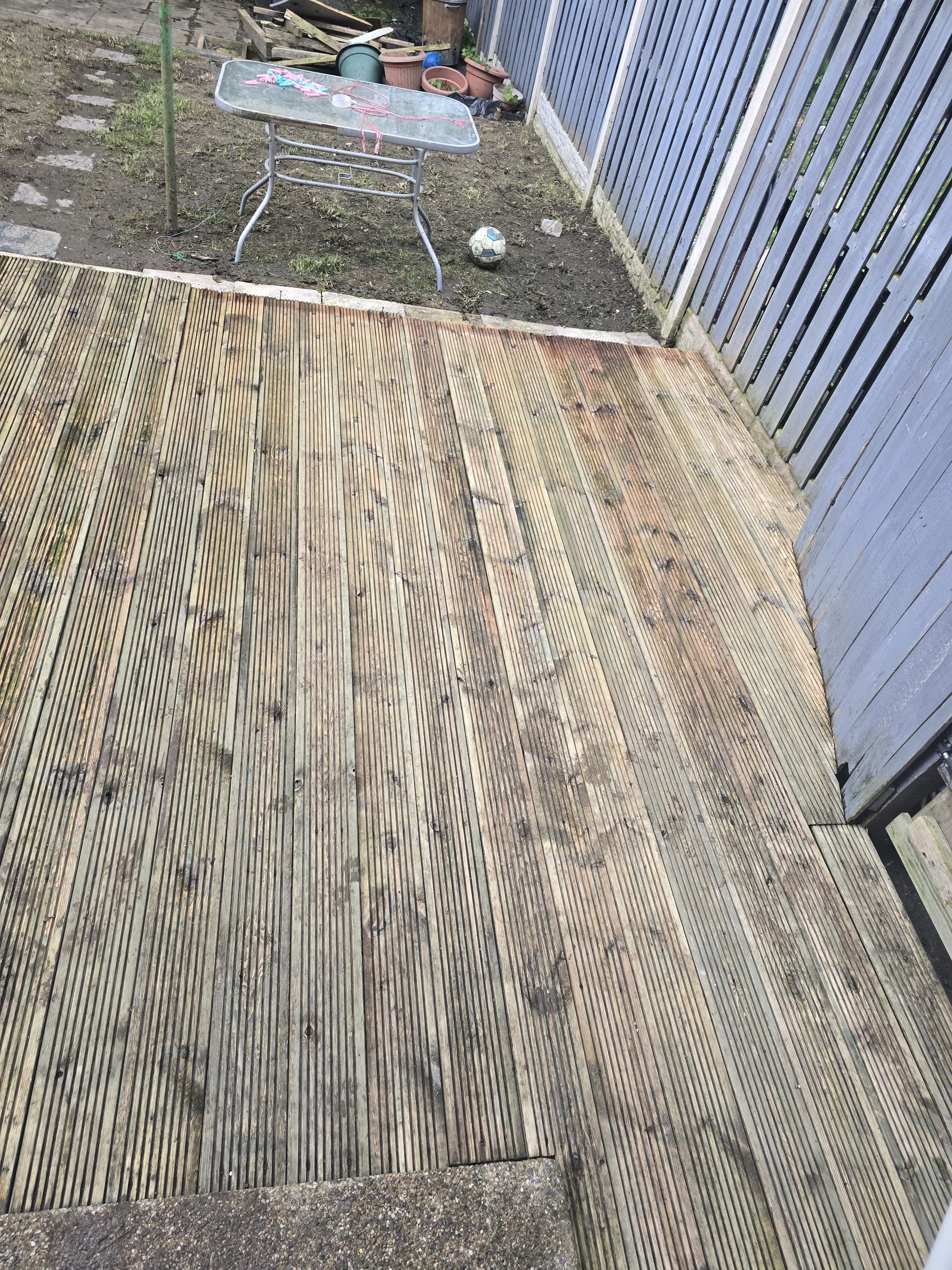 Partially wooden deck with a section of dirt and a small grassy area, including a glass-top patio table, a soccer ball, and various gardening pots and supplies against a wooden fence.