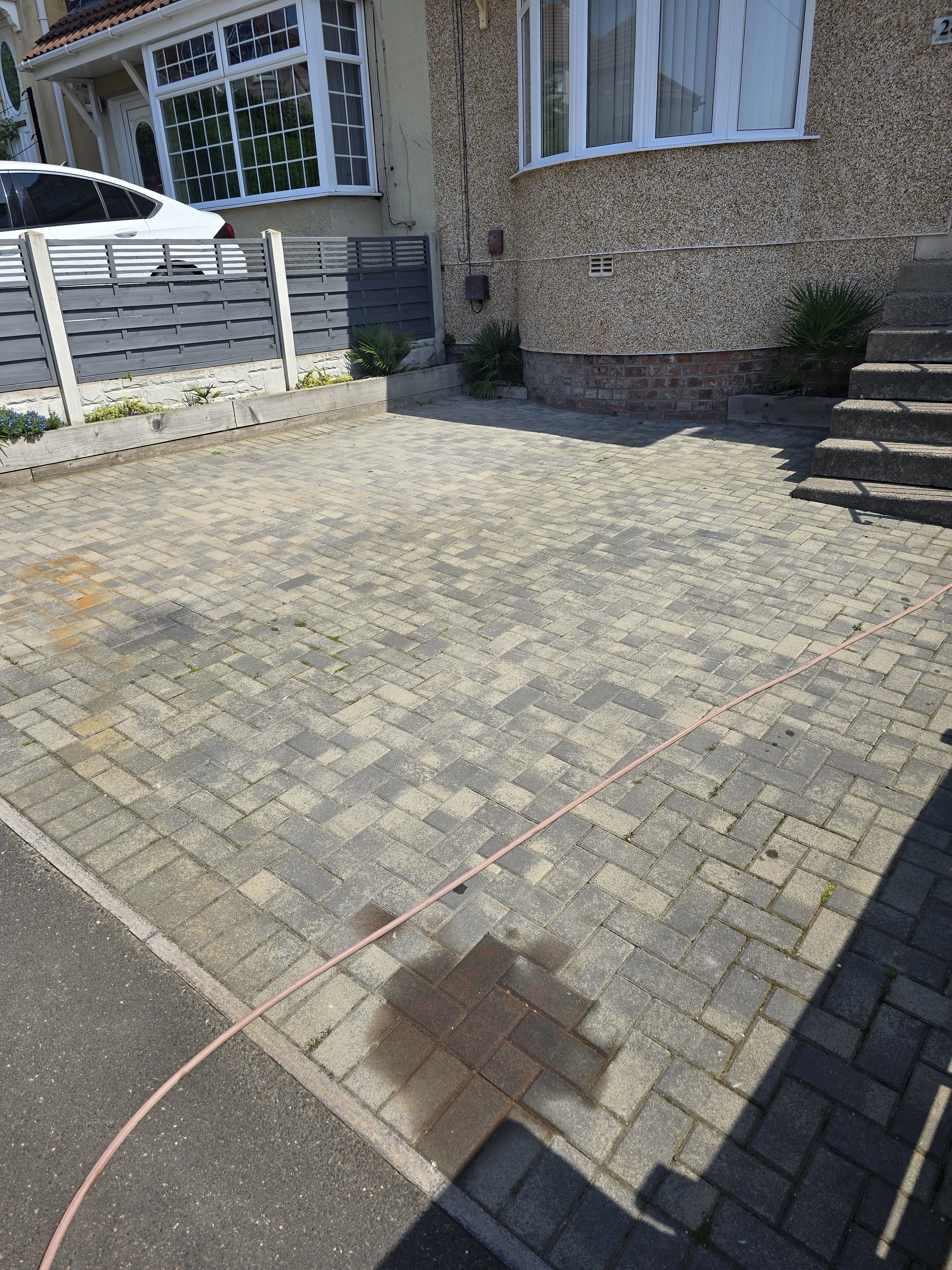A driveway with grey brick pavers and a sidewalk, part of a house with beige walls, a front window, a staircase, and a small garden bed with some plants.