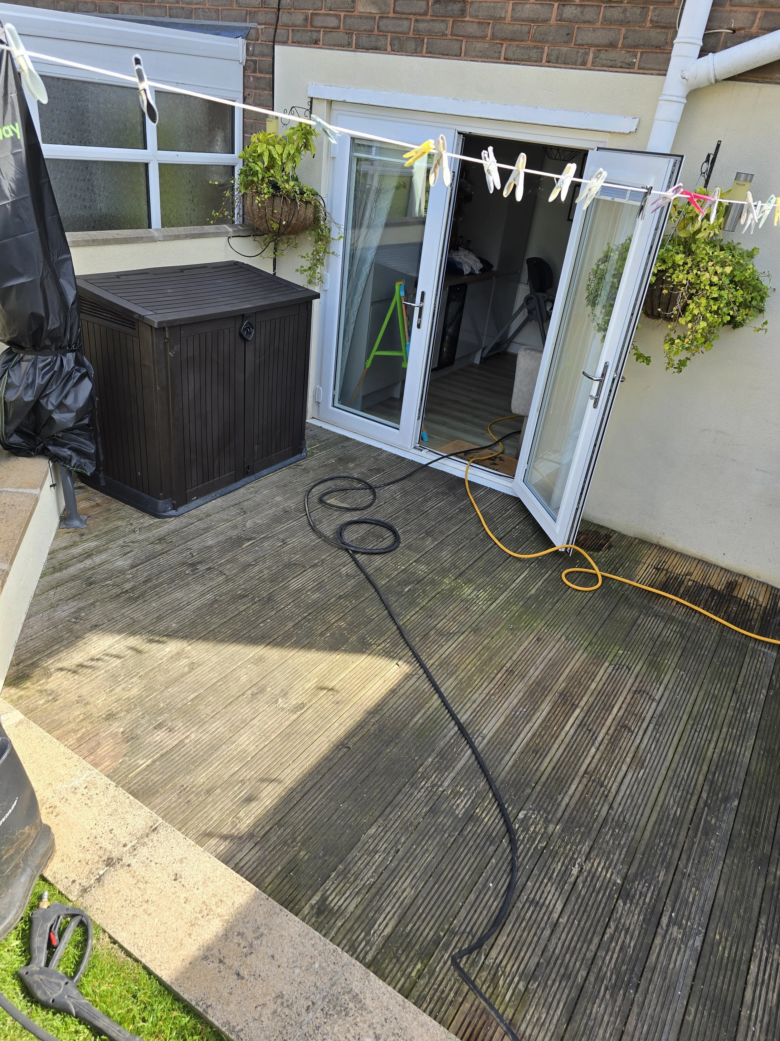 A patio with wooden decking, two hanging flower baskets, a black storage box, a clothesline with clothespins, a garden saw on the grass, and an open glass door leading to an indoor room.