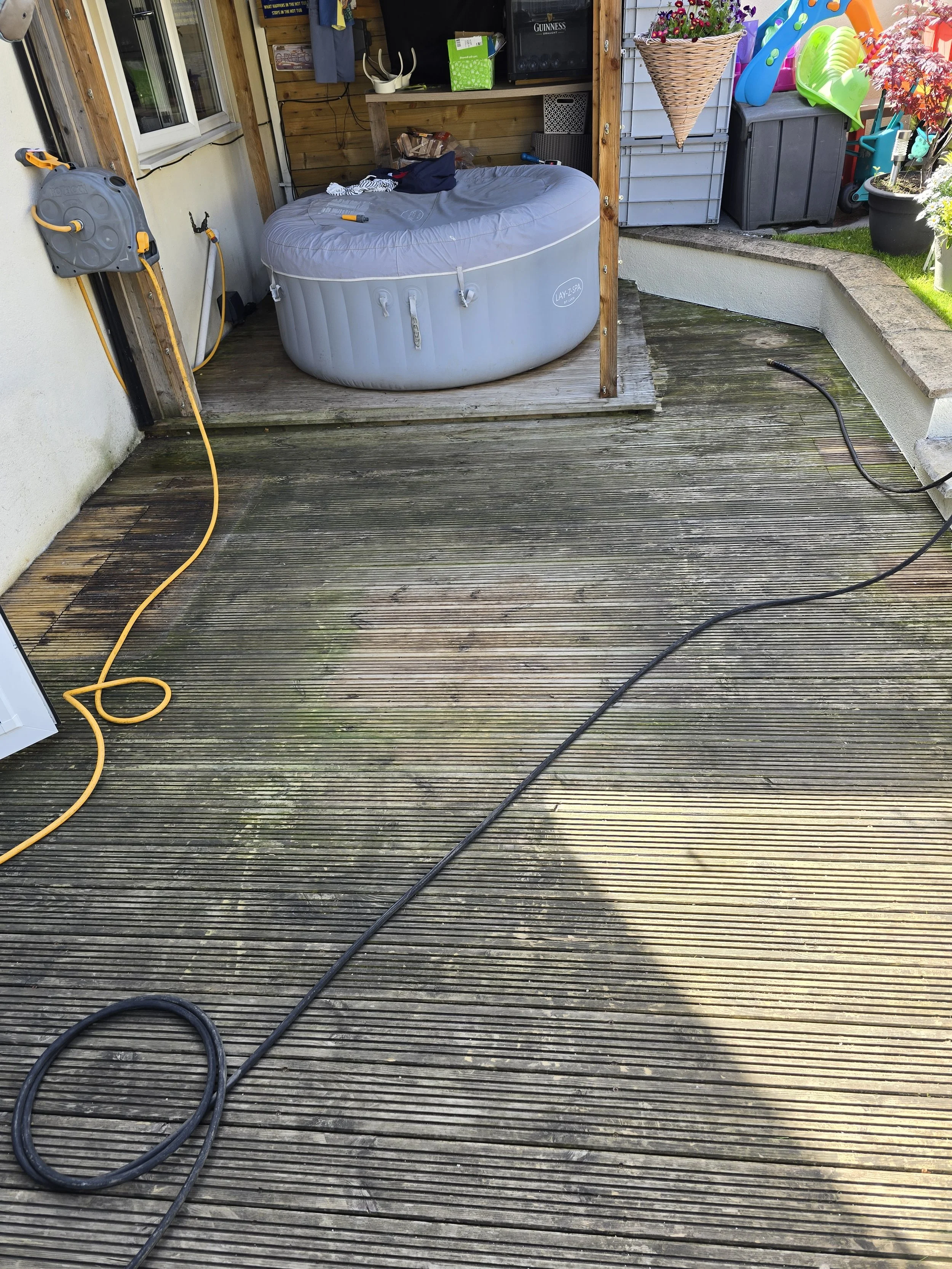 An outdoor wooden deck with a gray inflatable hot tub on the left side, connected to a hose reel on the wall. There are black and yellow extension cords on the deck. In the background, there are outdoor storage containers, gardening tools, and colorf
