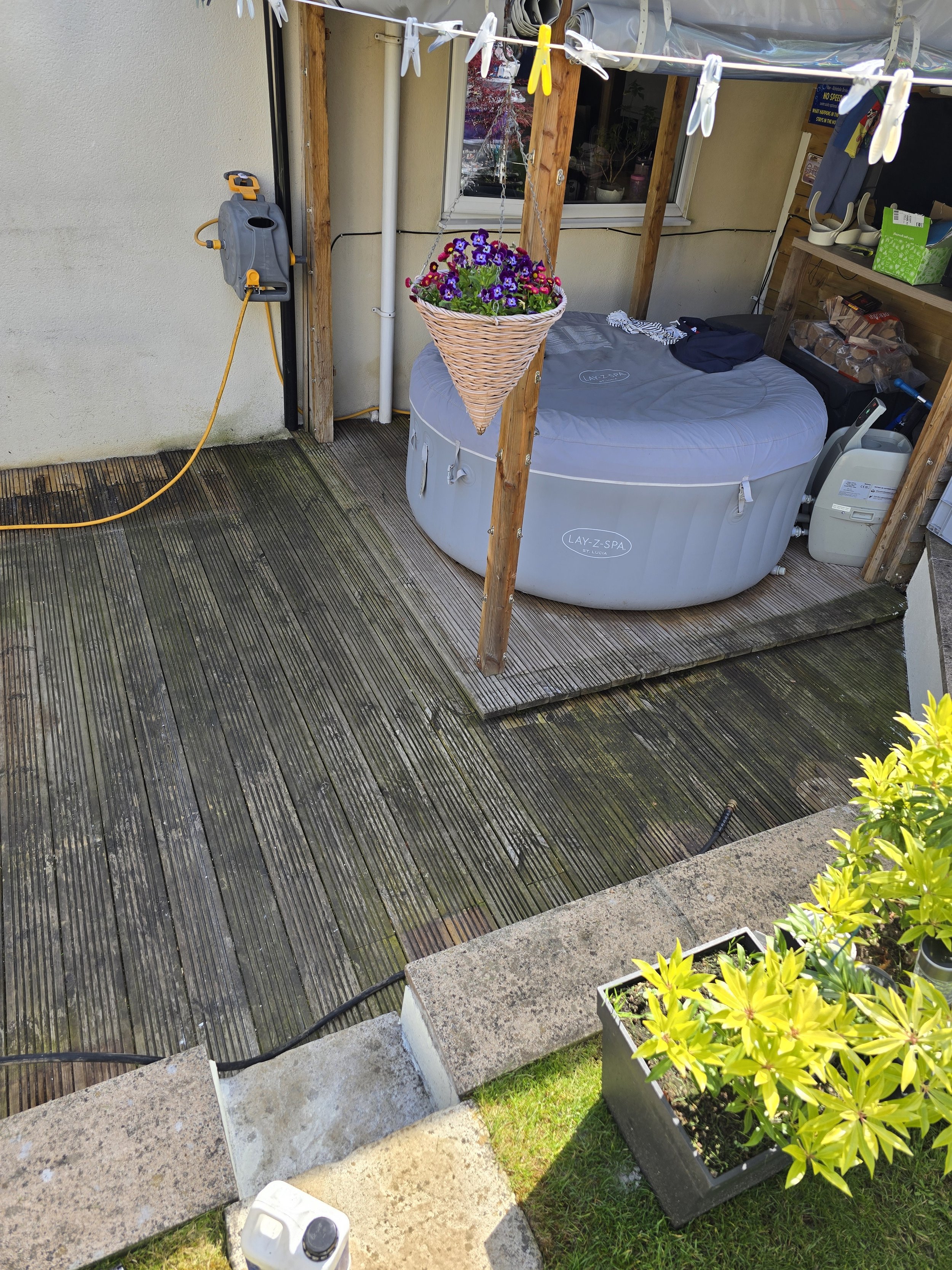 An outdoor patio area with a wooden deck, a gray hot tub, a flower basket hanging from a clothesline, and various gardening and household items placed on shelves and the ground, including a watering can, potted plants, and tools.