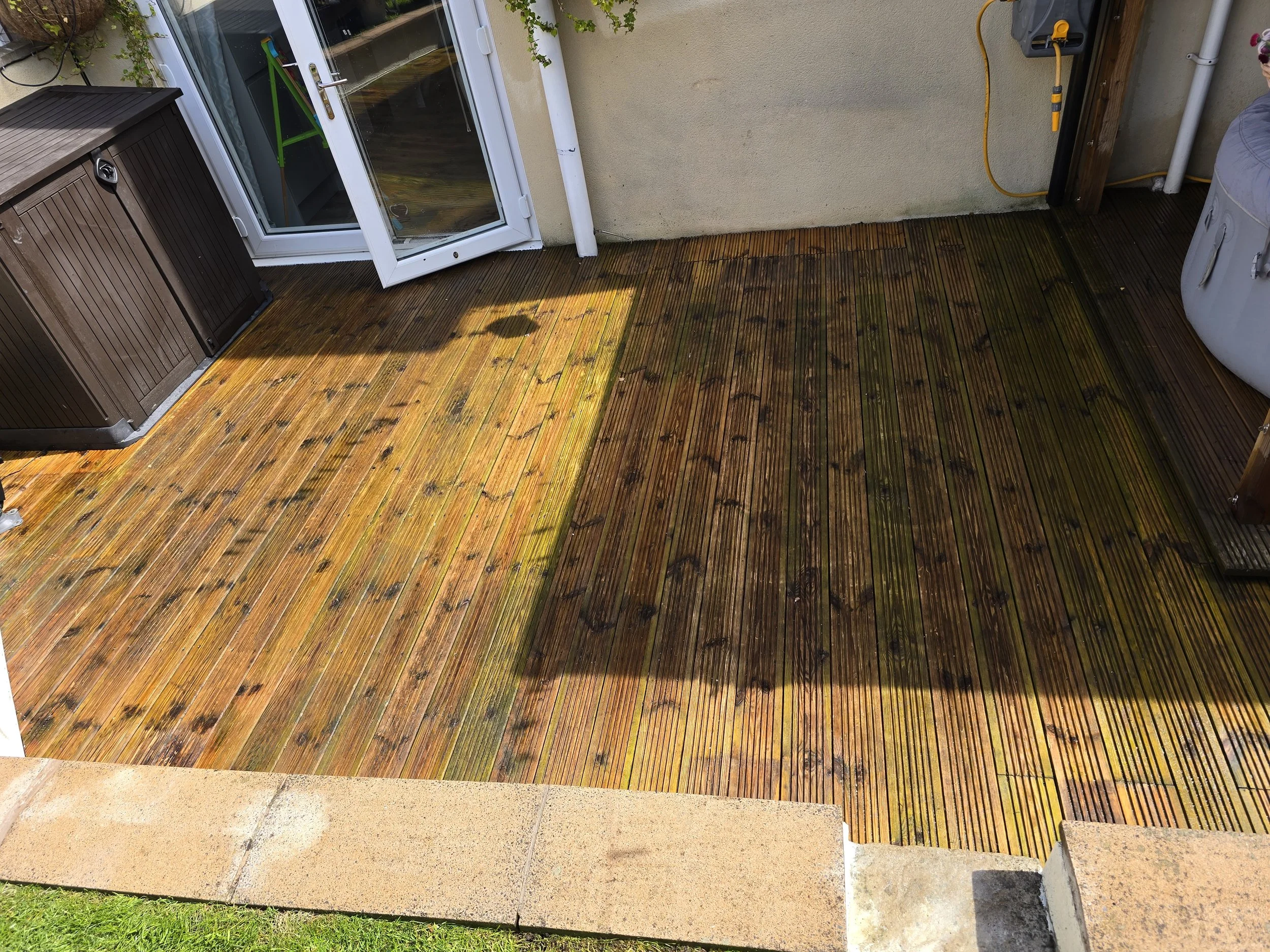 A wooden deck with a concrete border, a brown storage shed, a sliding glass door, a white drain pipe, and an outdoor washing machine. There are green plants and a yellow extension cord on the wall.