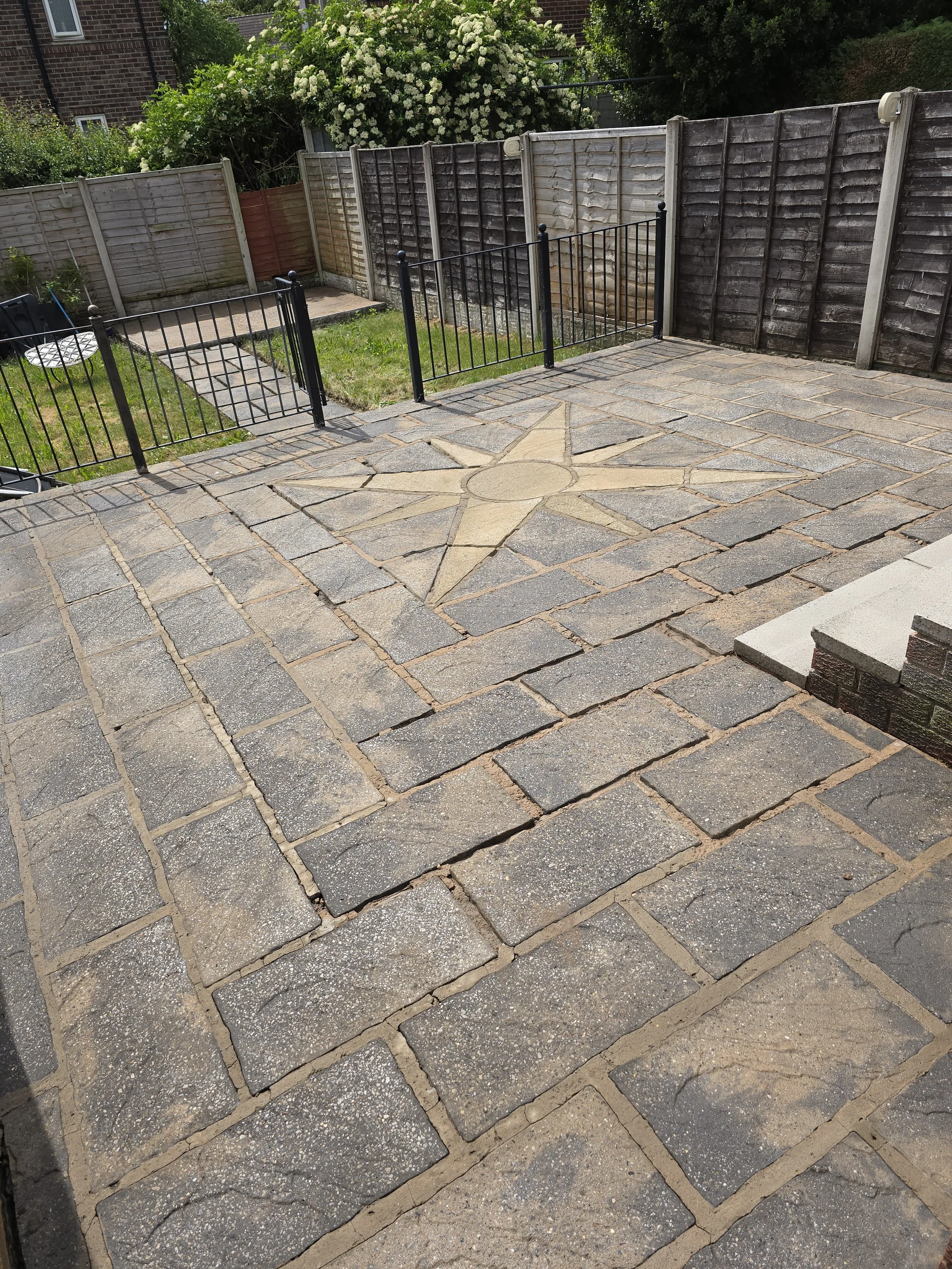 A backyard patio with stone paving forming a star-shaped design in the center, surrounded by bricks. There's a small grassy area, a black metal gate, and a wooden fence in the background with some greenery and bushes.