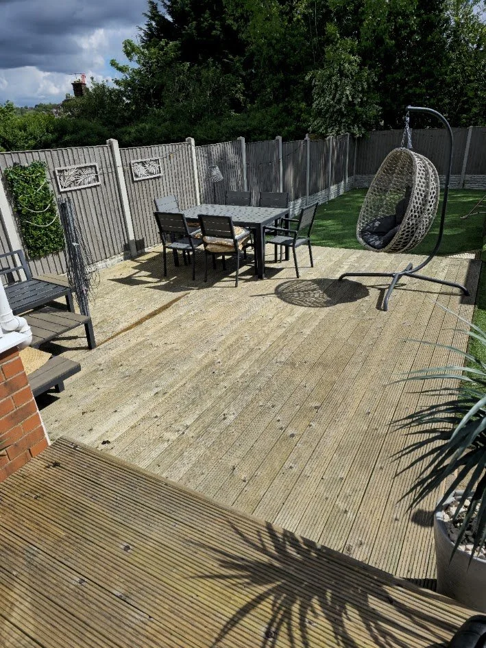 A backyard patio with a wooden deck, outdoor dining table with six chairs, a hanging egg chair, and a privacy fence surrounded by trees.