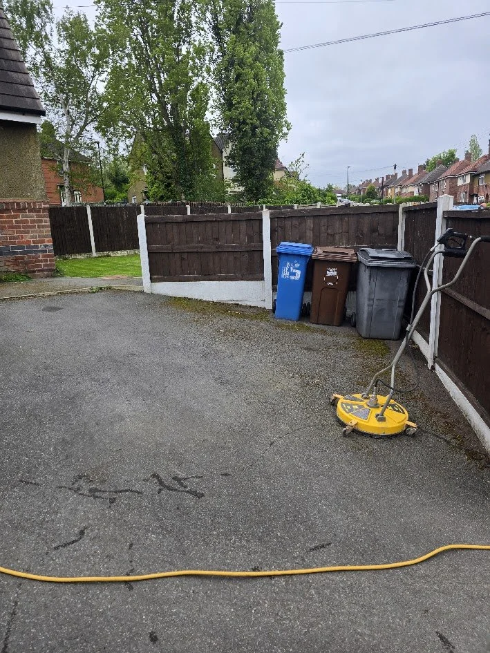 Backyard with garbage bins, a grass lawn, a wooden fence, and a yellow cleaning device on wet pavement.