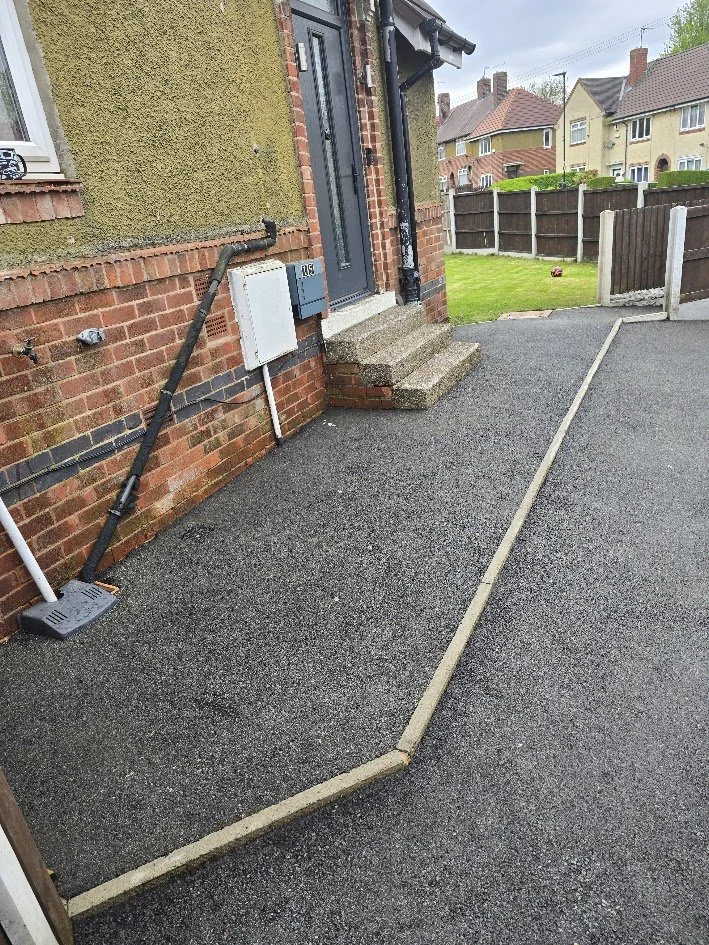 A paved driveway with a zigzag border marked by yellow paving stones, leading to the entrance of a house with concrete steps. The house has a brick exterior and a blue door, with a black drainpipe running down the wall. There is a green lawn enclosed