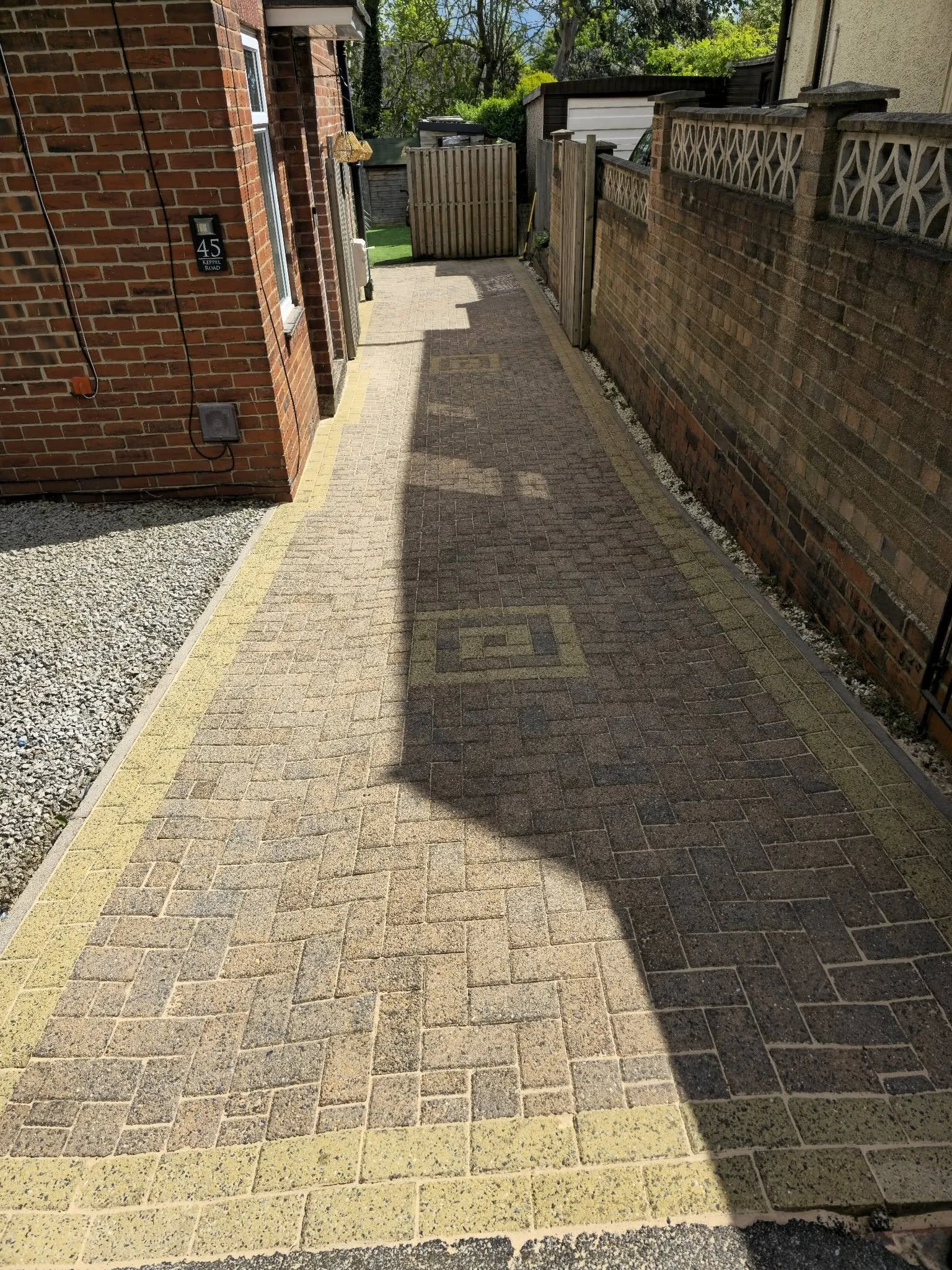 A small, paved walkway runs between two brick walls, leading to a gate in the background. The walkway is bordered by gravel on one side and a yellow border on the other.
